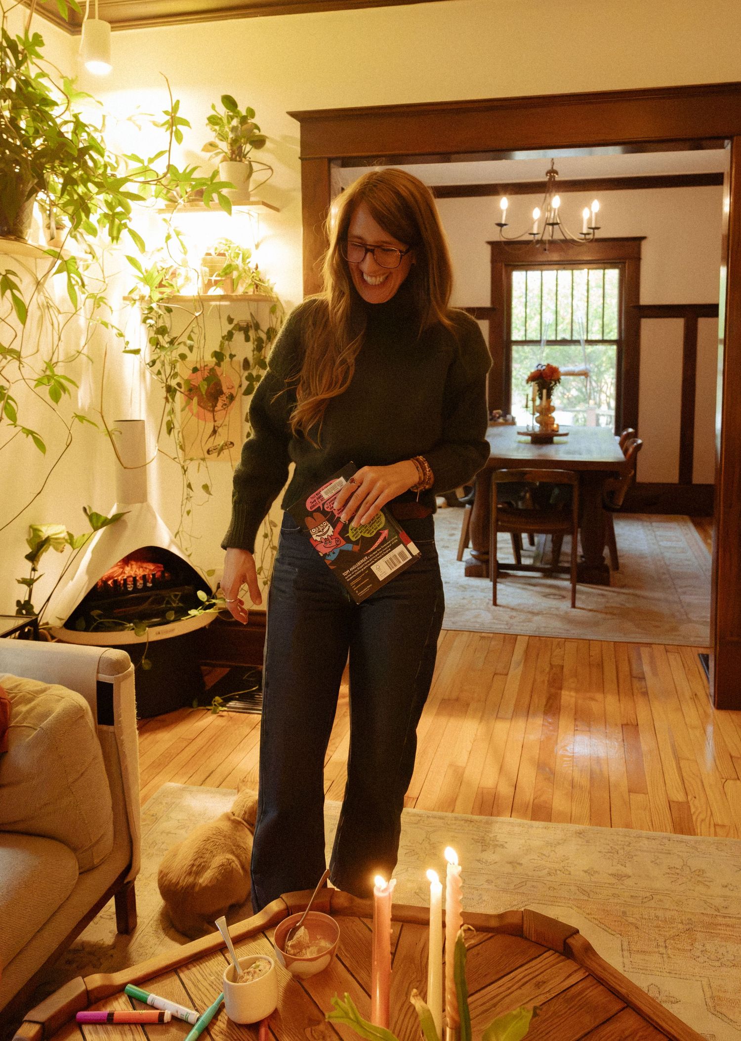 Smiling woman in glasses holds a book in a cozy living room with hanging plants, a lit fireplace, candles, and a wooden dining table.