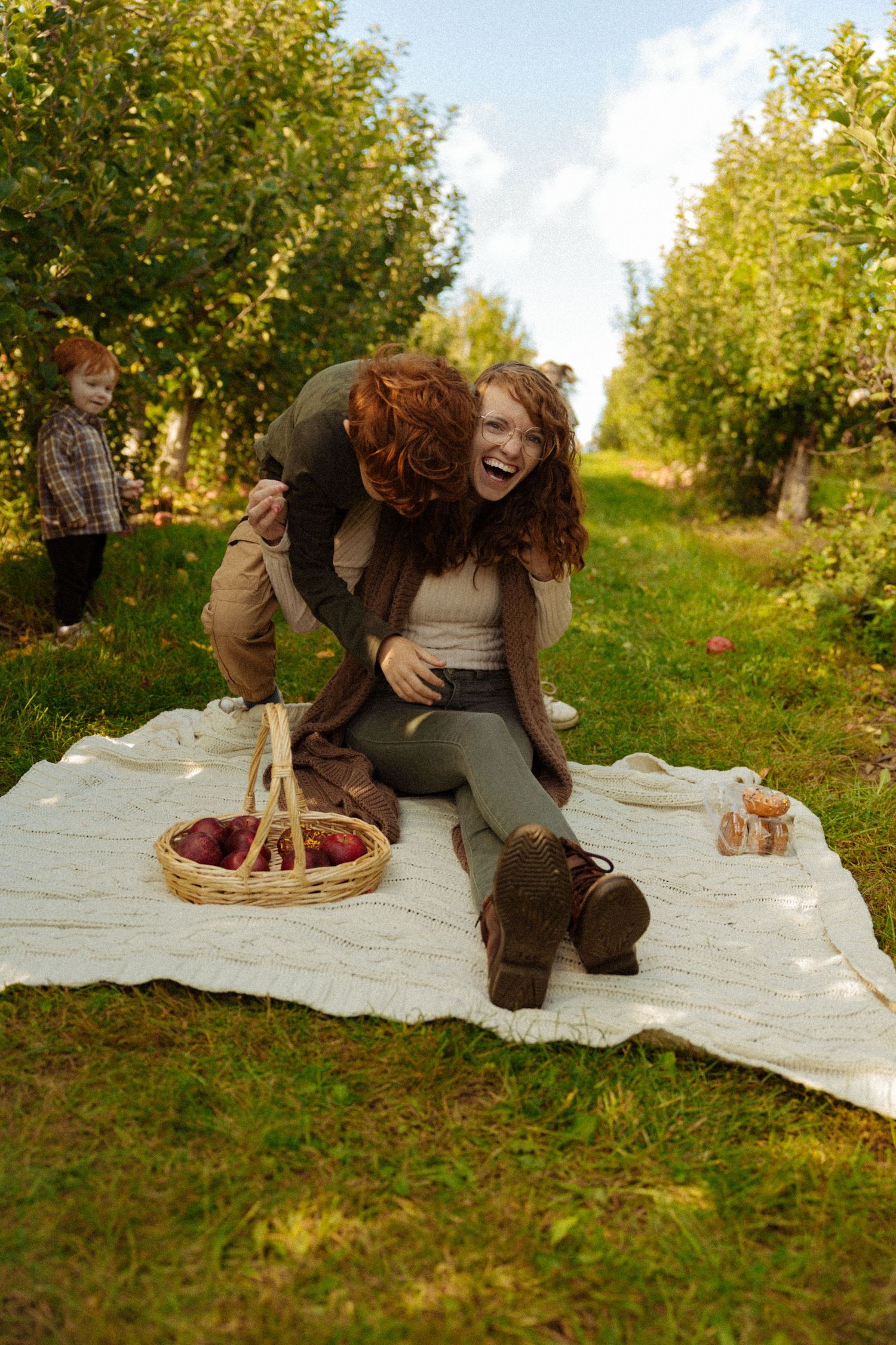 Couple laughing on a picnic blanket in an apple orchard with a basket of apples and a child in the background.