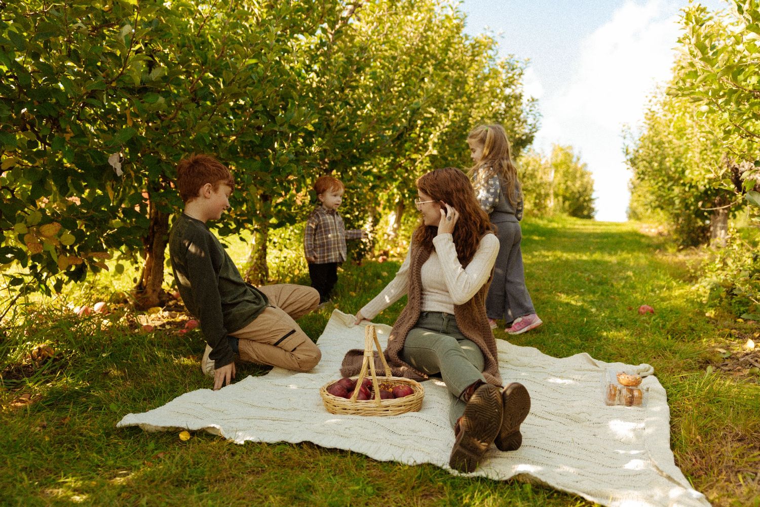 Family enjoying a picnic on a white blanket in a lush green orchard on a sunny day.