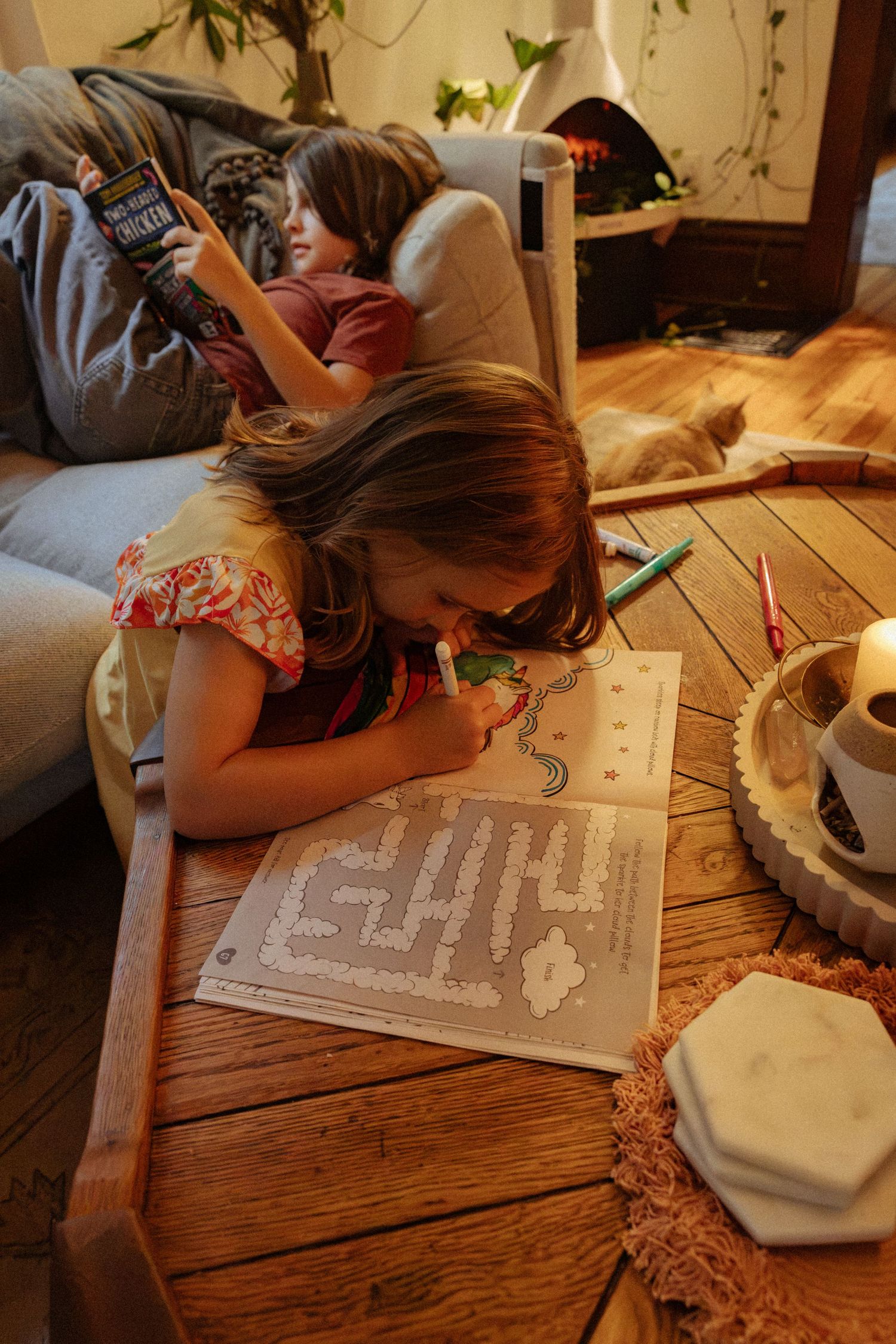 A young girl colors a maze activity page at a wooden coffee table while a child reads on the couch near a cozy fireplace.