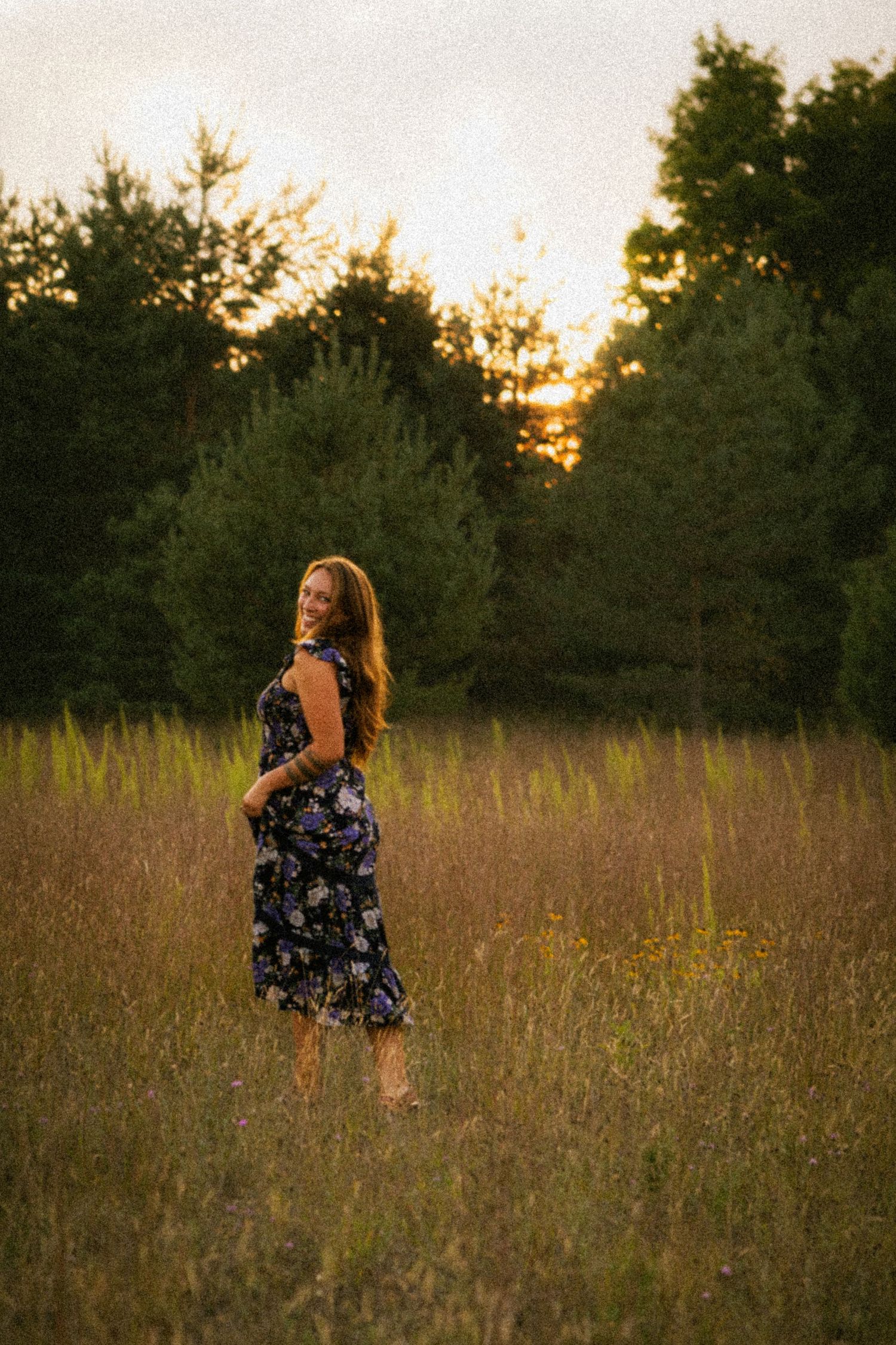 Woman in floral dress standing in golden field at sunset with green trees in background.