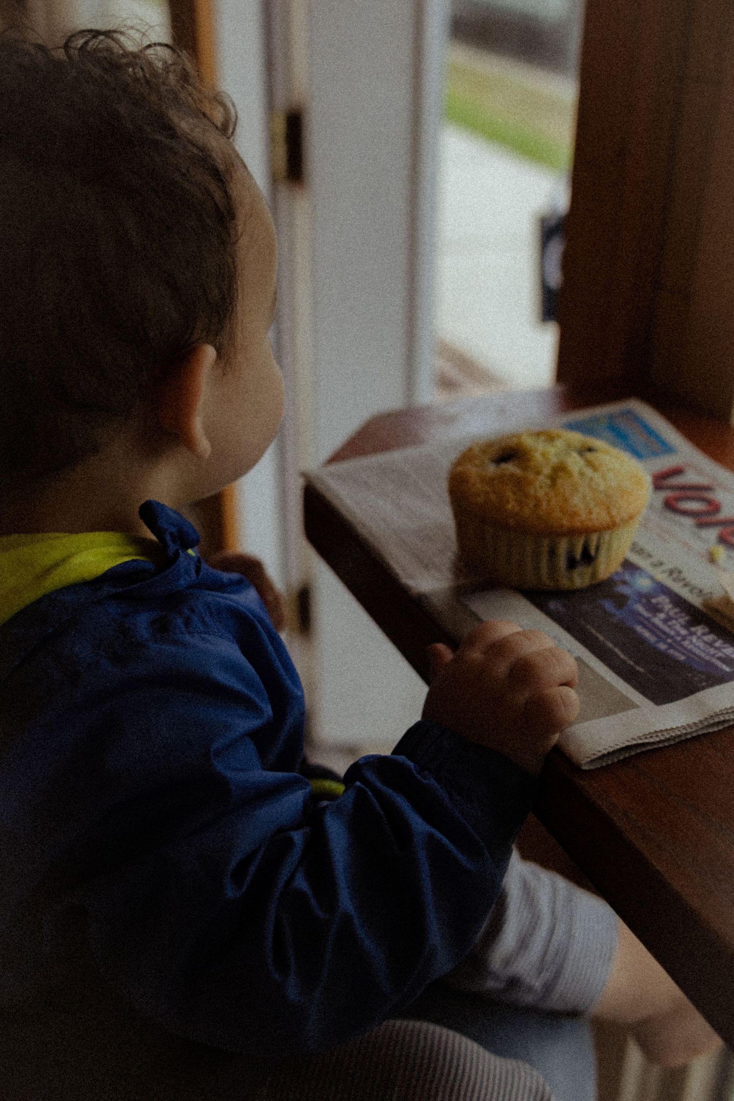 Young child sitting at a wooden table with a blueberry muffin on a newspaper.