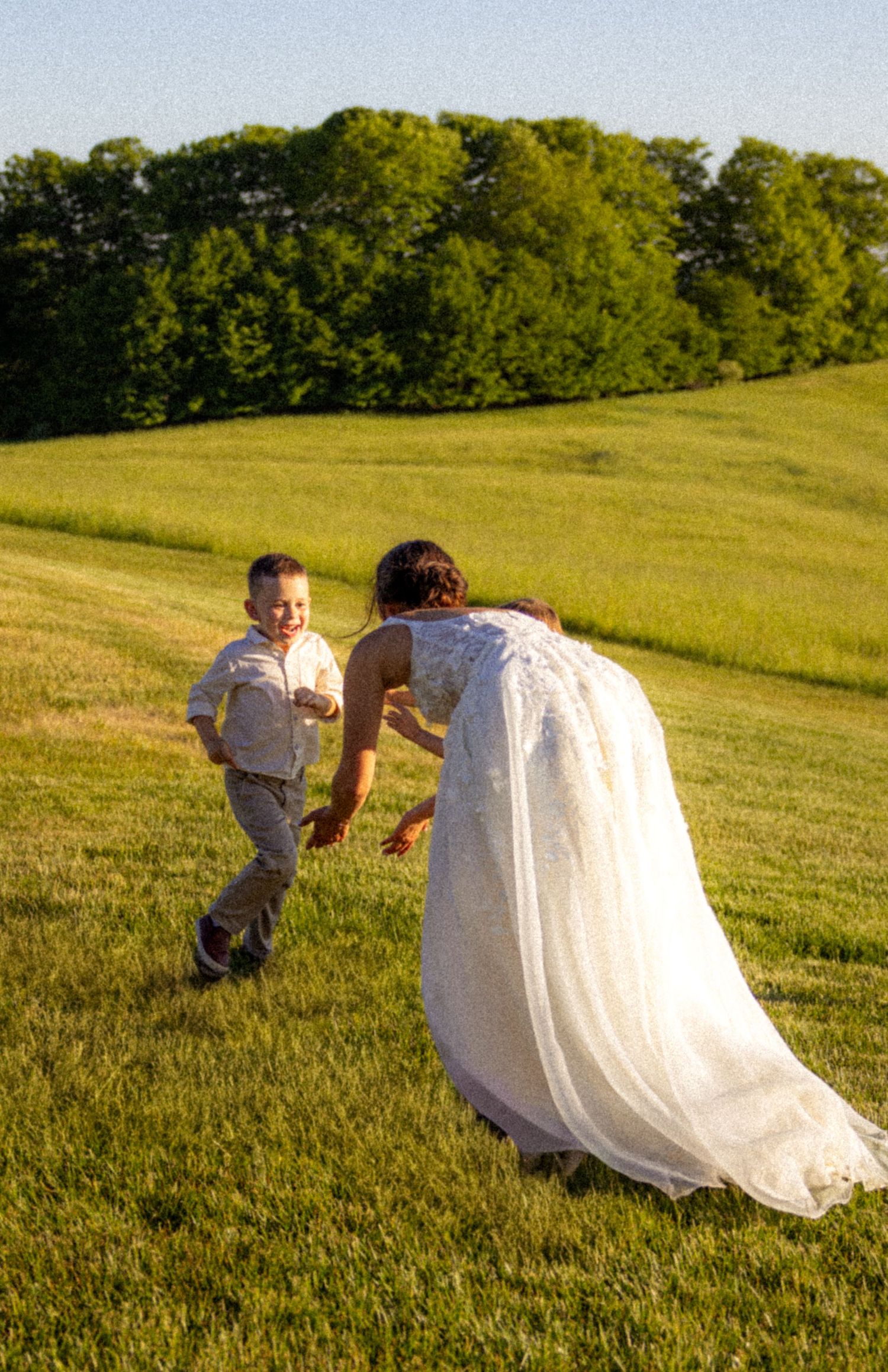 A bride in a white gown reaches toward a smiling young boy running across a sunny green field.