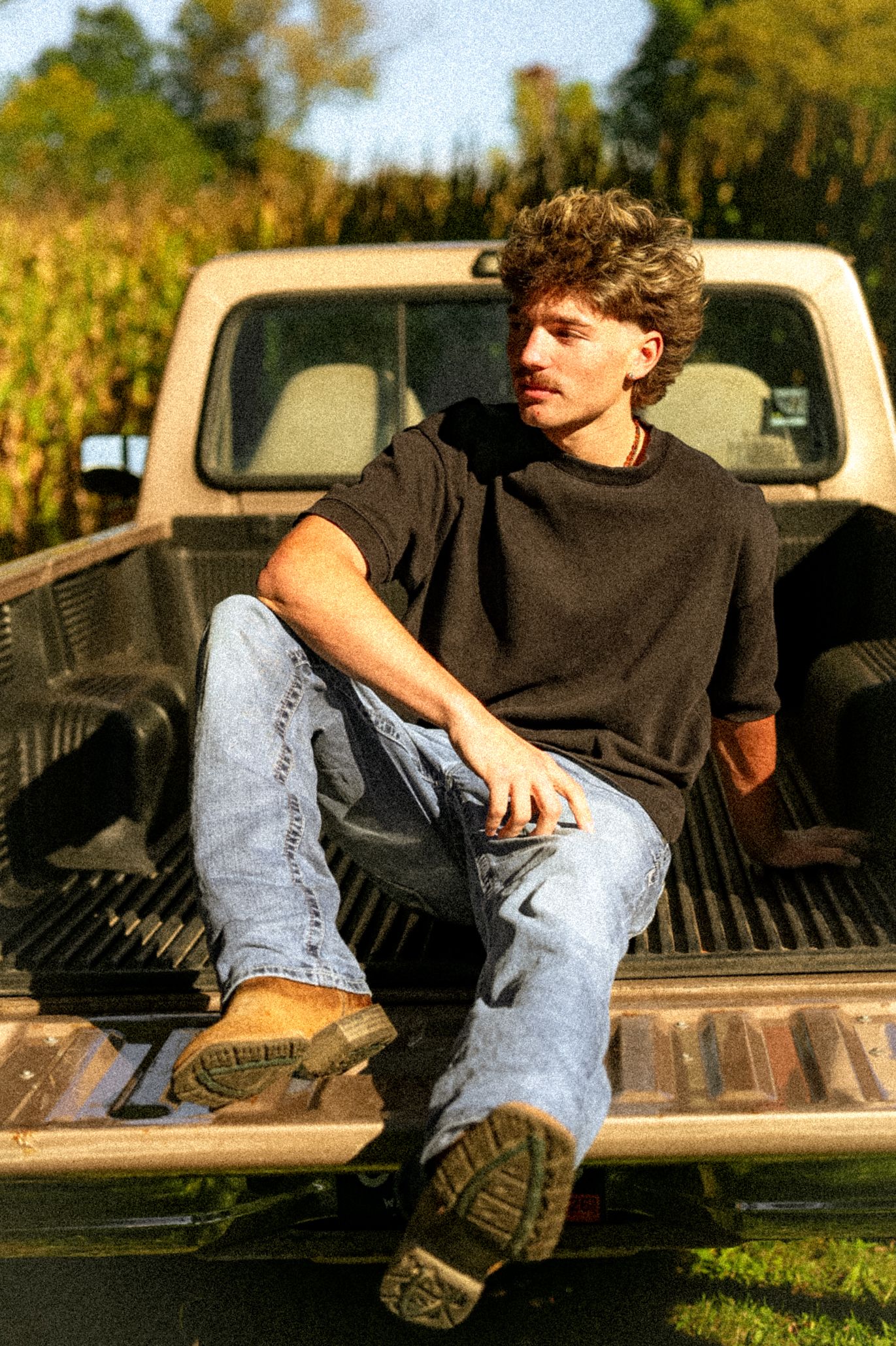 Young man in black shirt and jeans sitting on pickup truck bed near cornfield on sunny day.