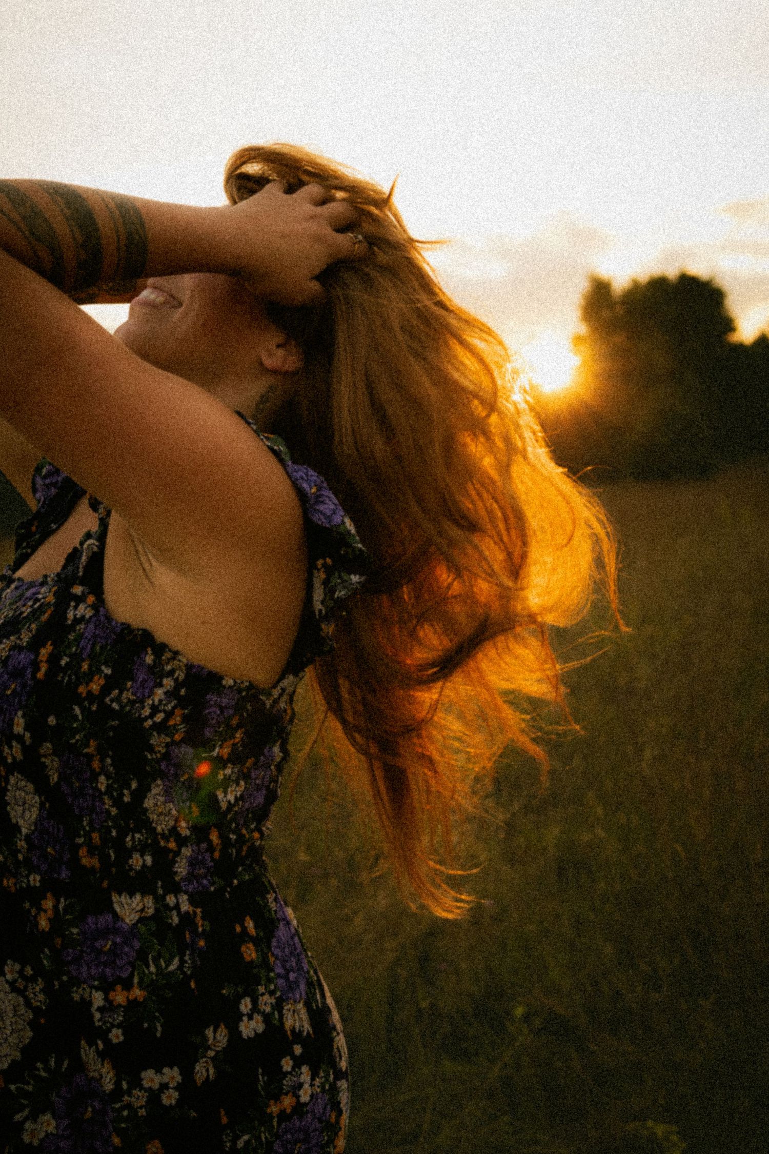 Woman in floral dress tosses long hair backlit by golden sunset in a green field.