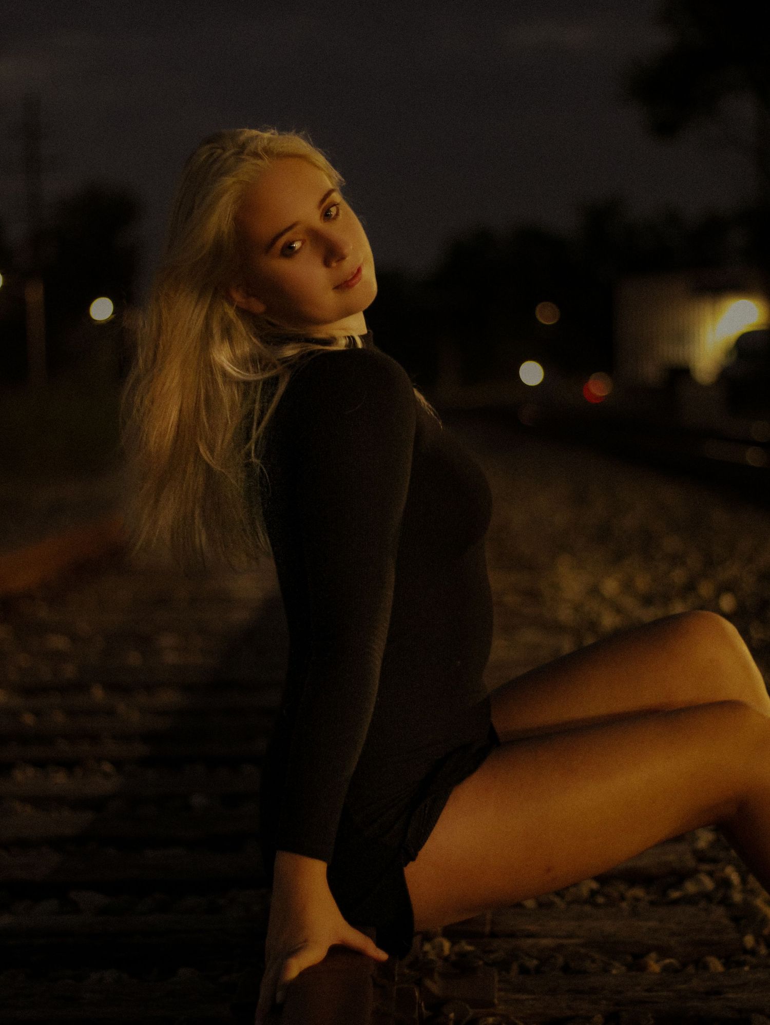Young blonde woman in black outfit sitting on railroad tracks at night with bokeh lights in background.
