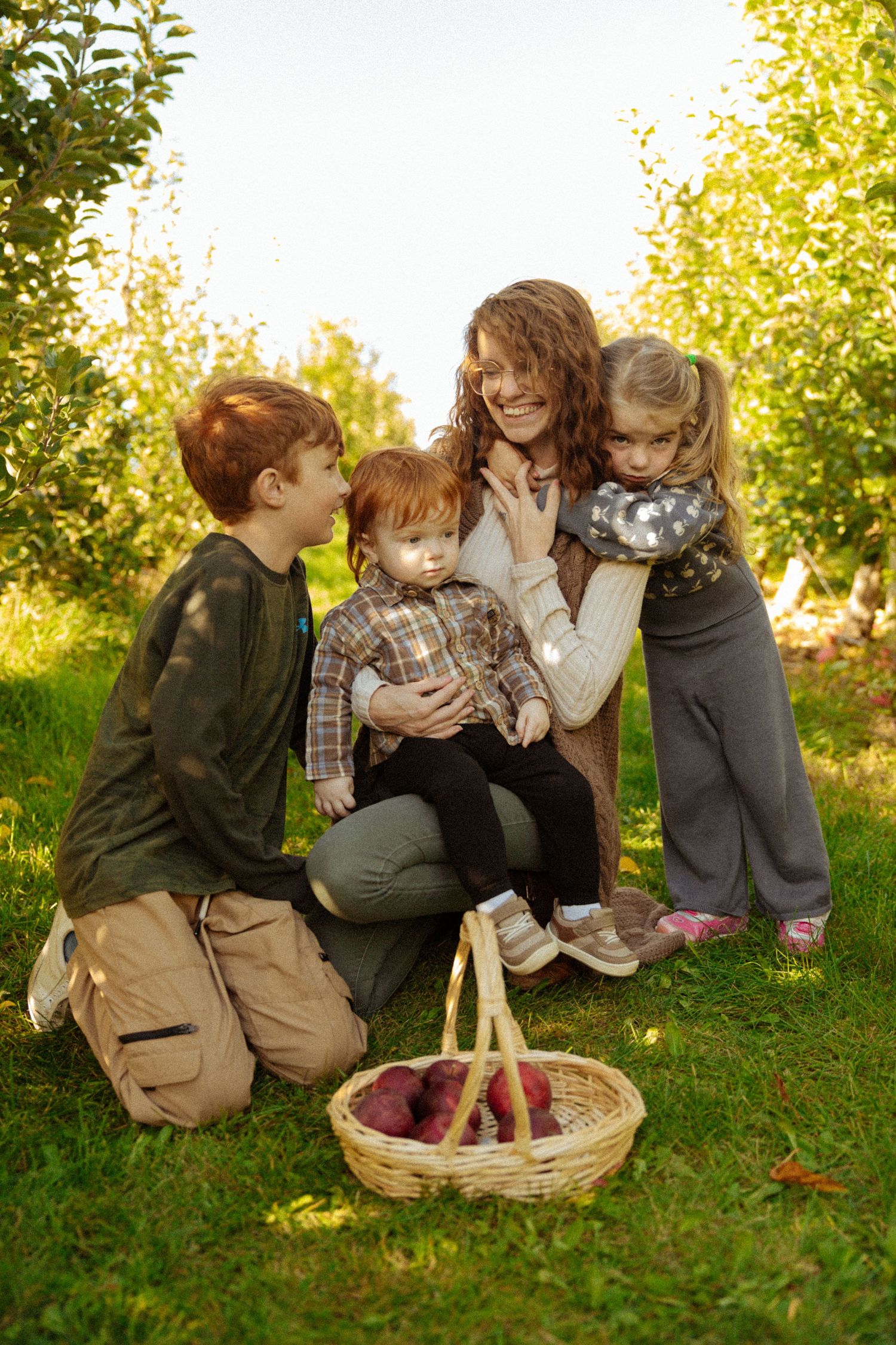 A smiling mother kneels on grass in an orchard with three children and a basket of red apples on a sunny day.