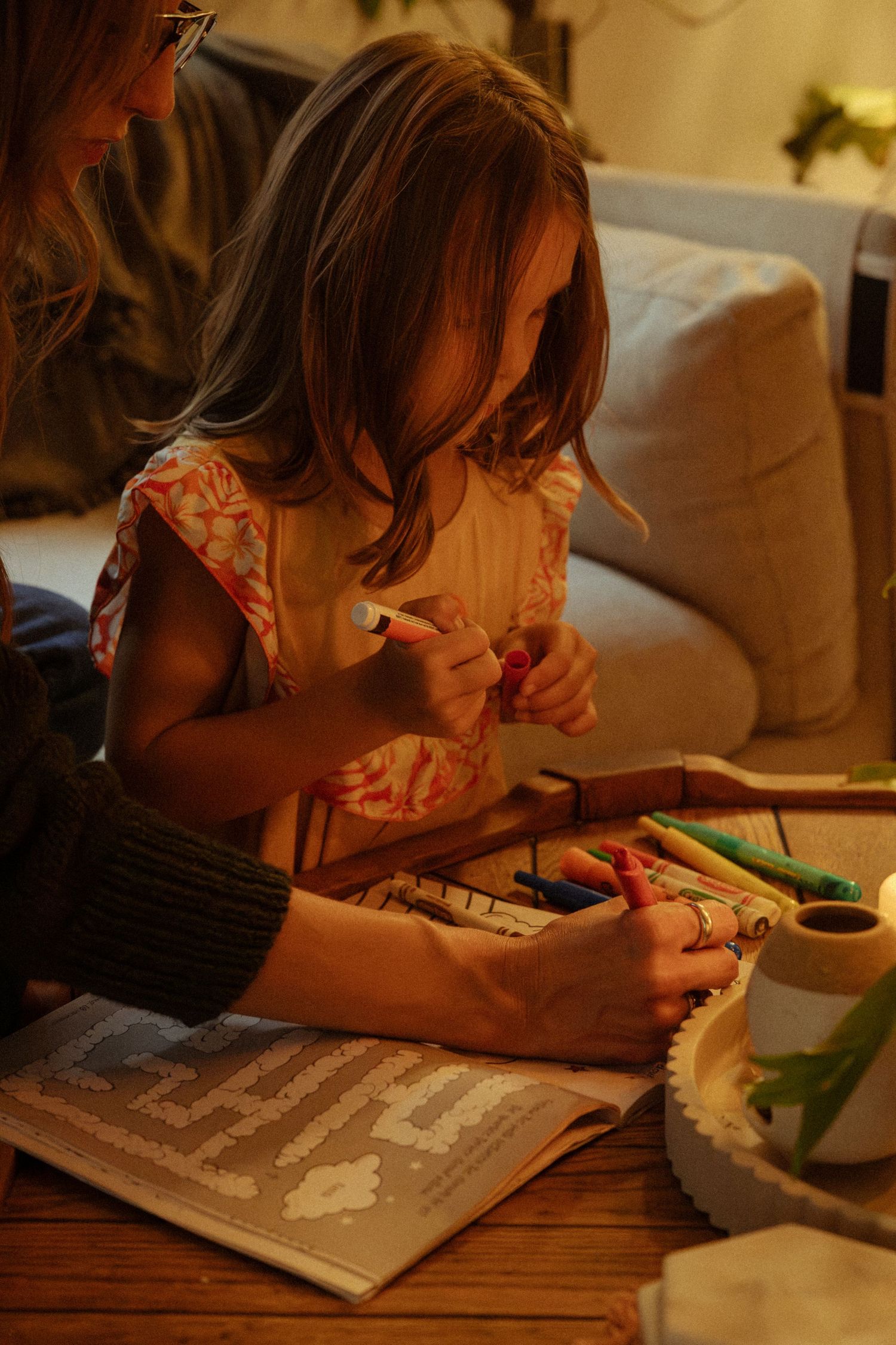 A girl in a floral top colors with markers at a wooden table while a woman helps with a puzzle book nearby.