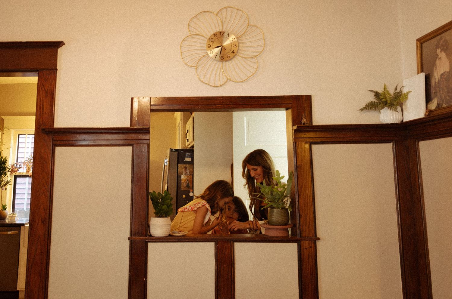 Two people sit close together in a cozy room, reflected in a wood-framed mirror beneath a gold sunburst wall clock and potted plants.
