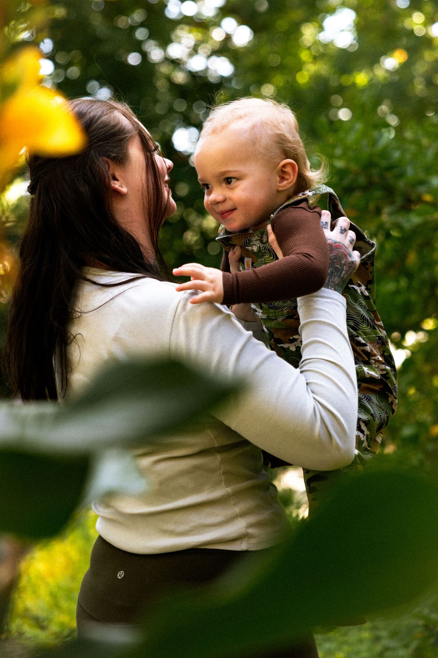 A mother lovingly holds her smiling baby in a camouflage outfit outdoors among green trees.