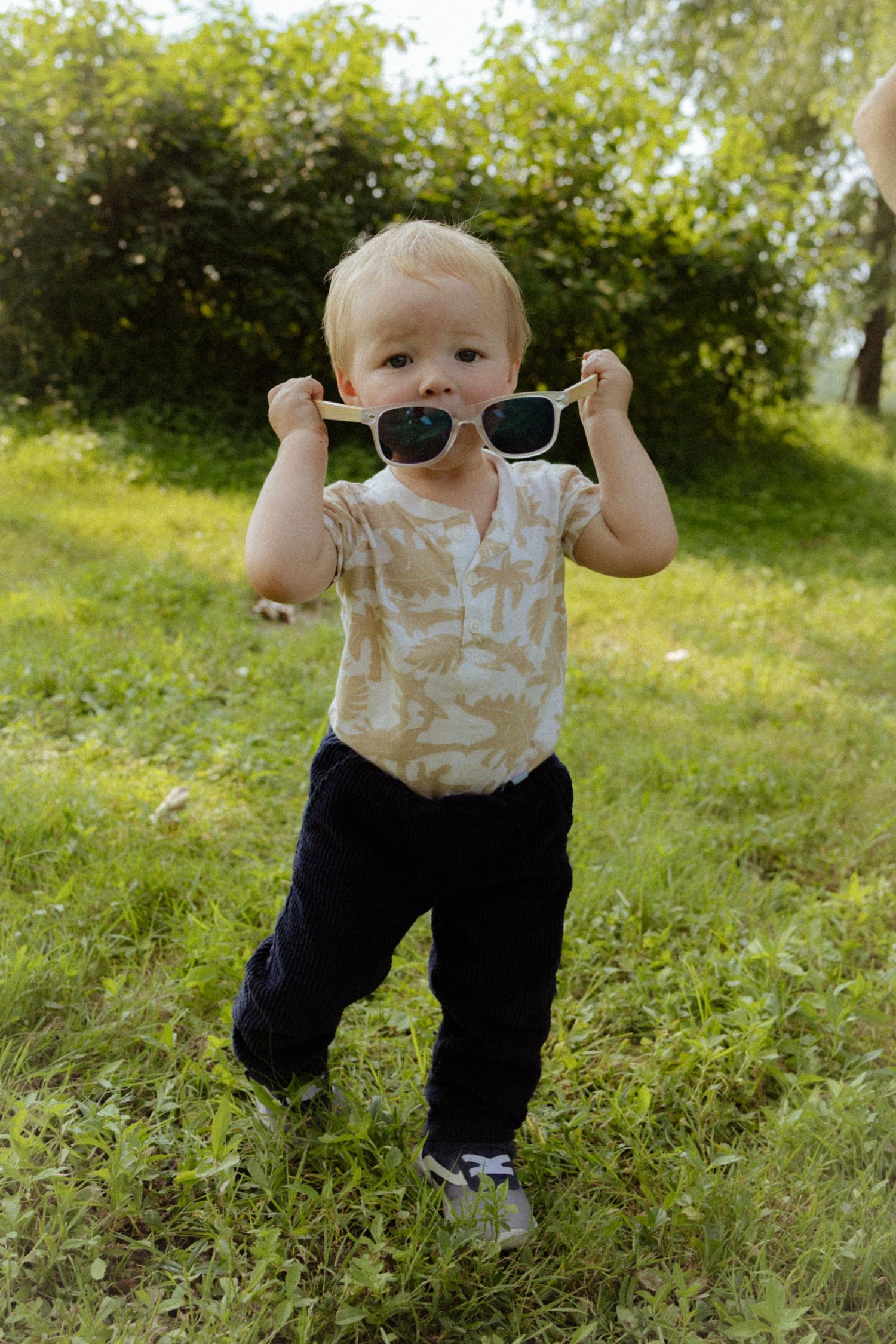 A cheerful blonde toddler boy chewing on white sunglasses while standing on a green grassy lawn.