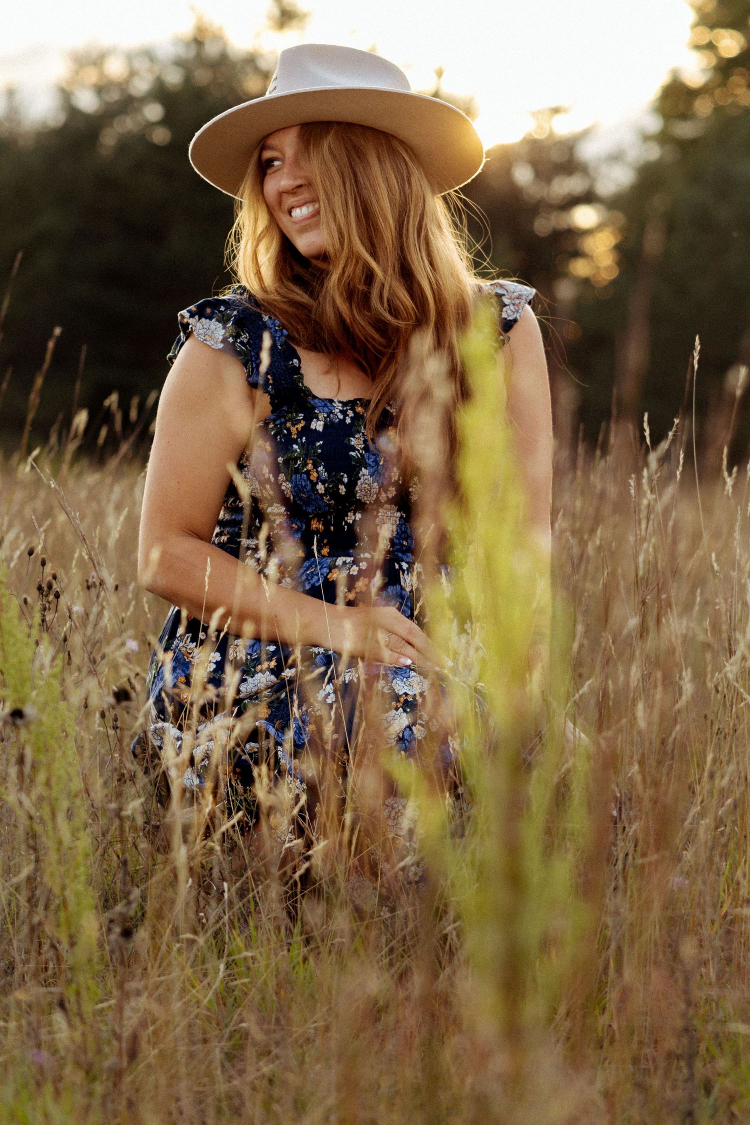 Smiling woman in blue floral dress and white hat stands in golden sunlit meadow at sunset.