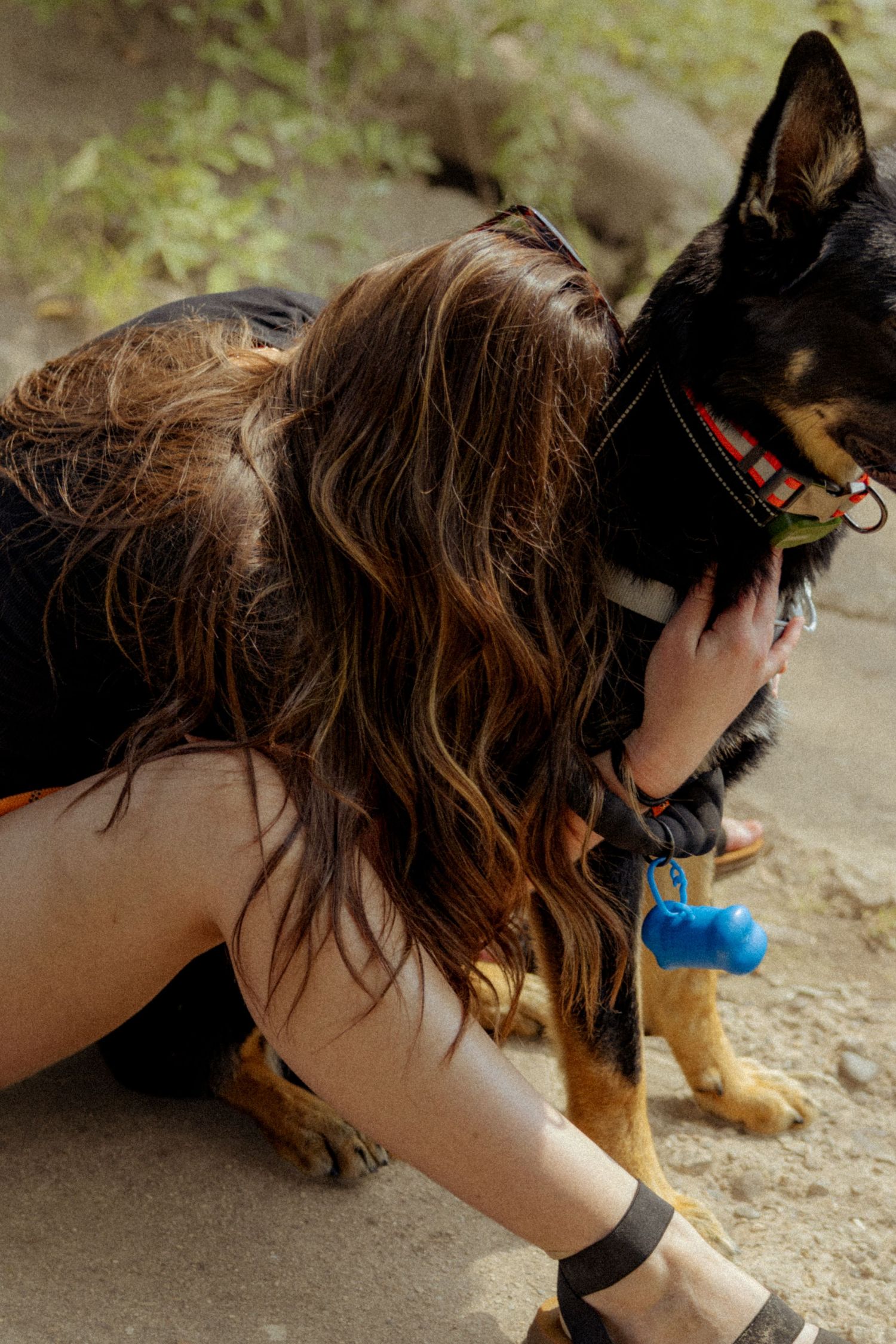 A woman with long brown hair hugs a black and tan German Shepherd dog wearing a red collar outdoors.