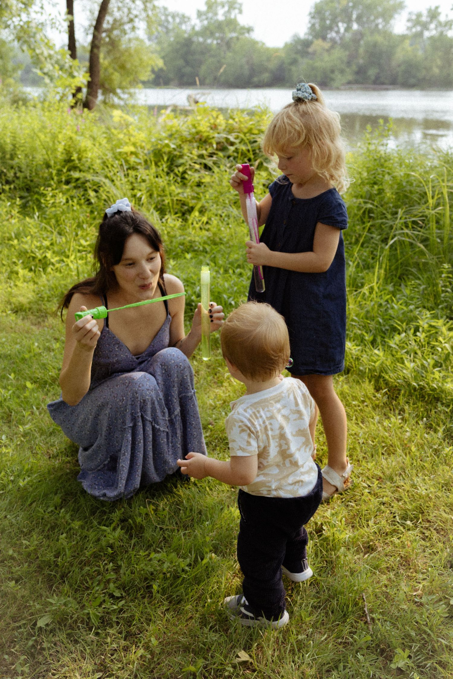 A mother blows bubbles with her two young children on a grassy riverbank surrounded by lush green trees.
