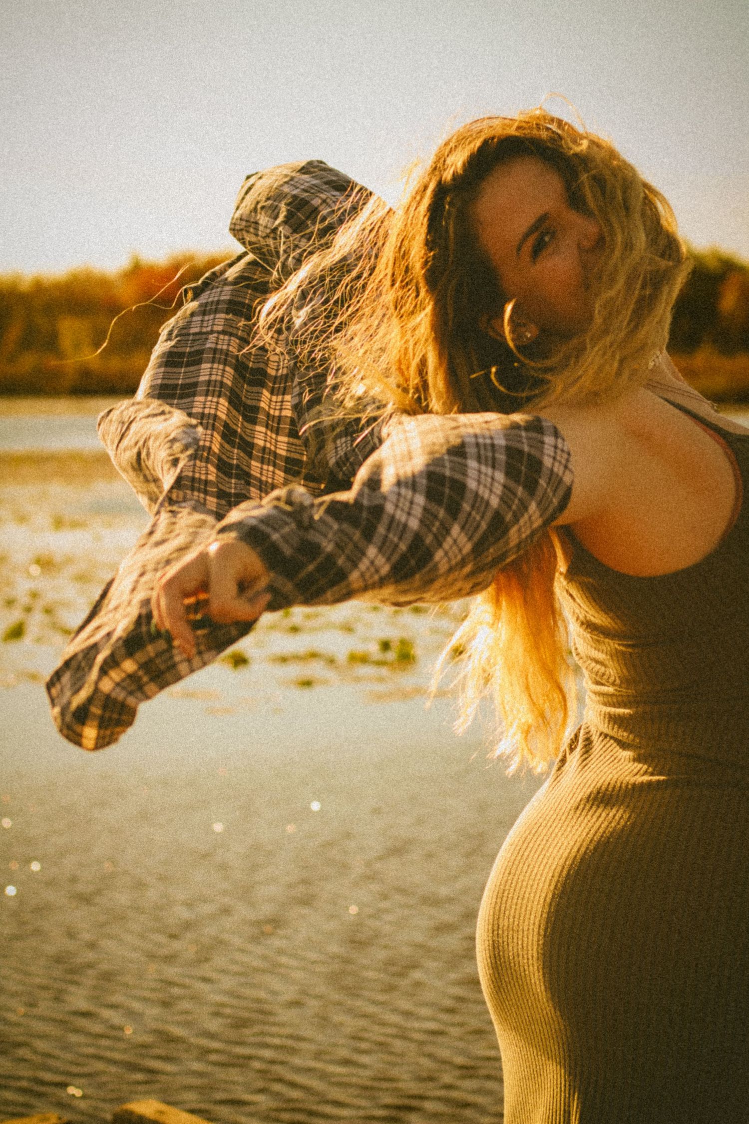 Woman in plaid shirt and olive dress poses by a sunlit lake, her long hair blowing in the wind at golden hour.