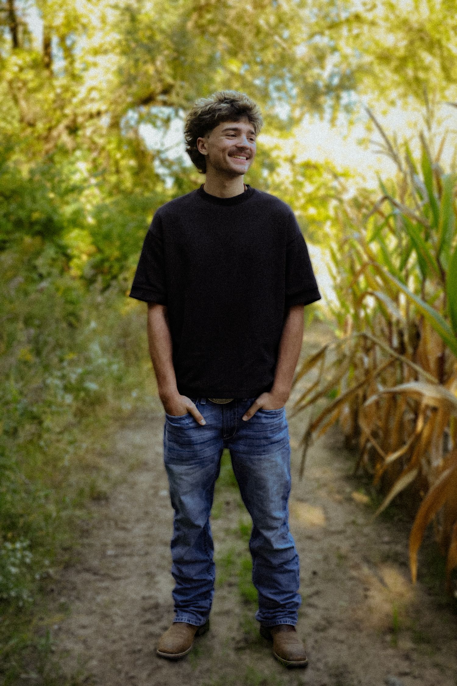 Young man in black t-shirt and jeans smiling outdoors on a dirt path surrounded by green trees and corn stalks.