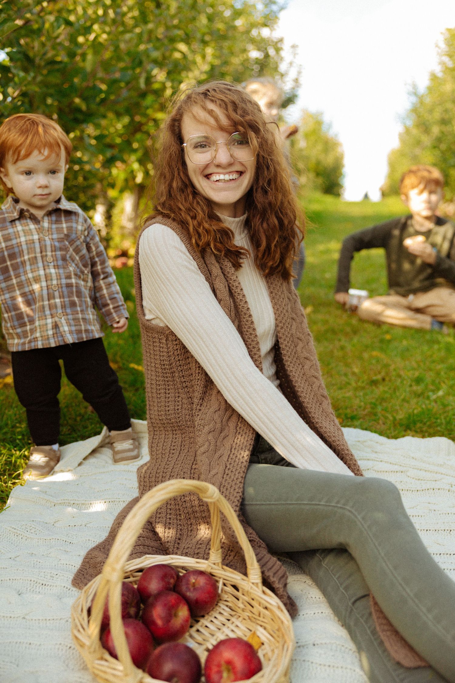 A smiling redhead woman sits on a blanket at a family picnic with a basket of red apples, children playing in background.