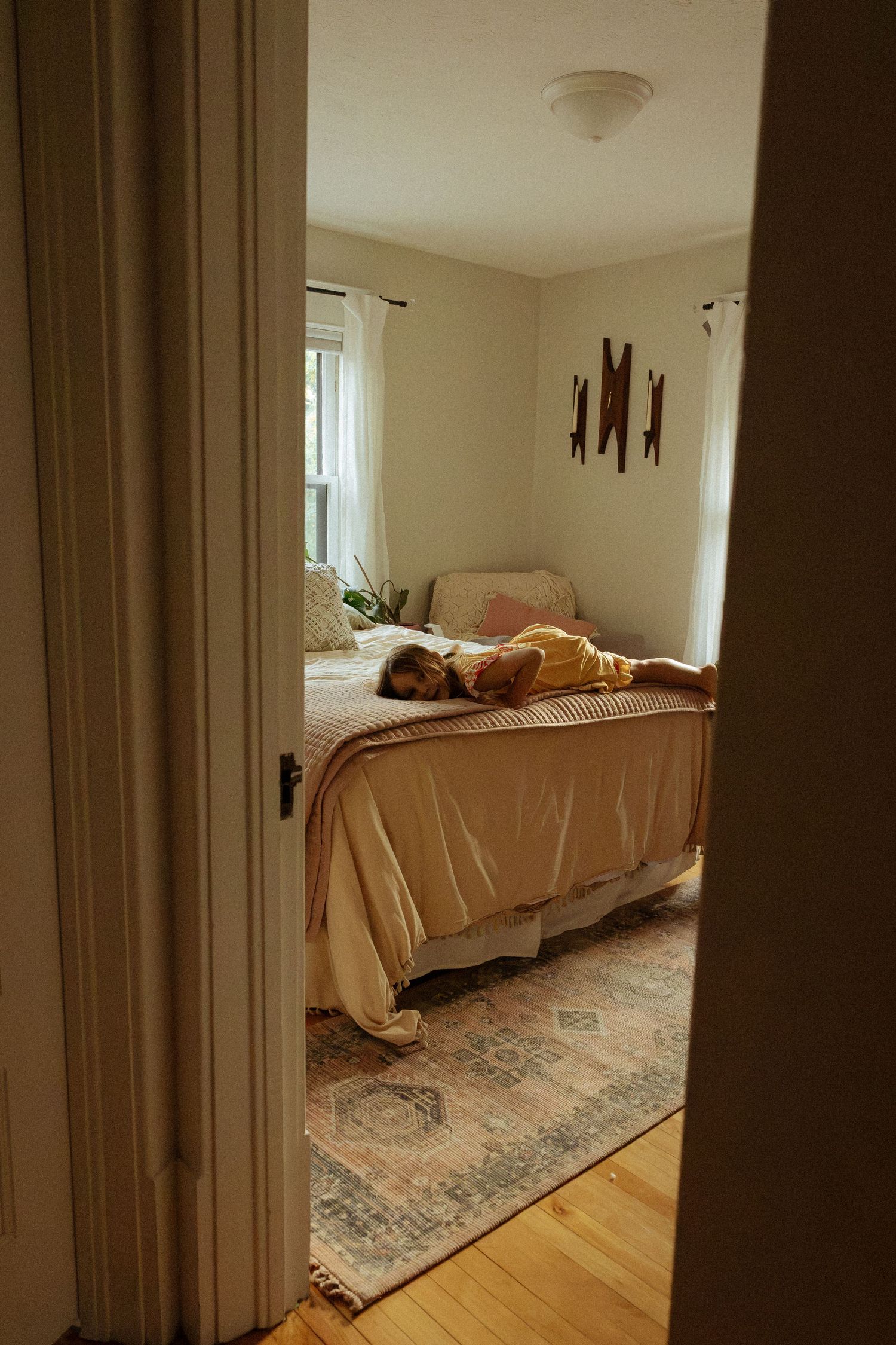 A child lies on a made bed with a beige coverlet in a sunlit bedroom, seen through a doorway, with a patterned rug below.