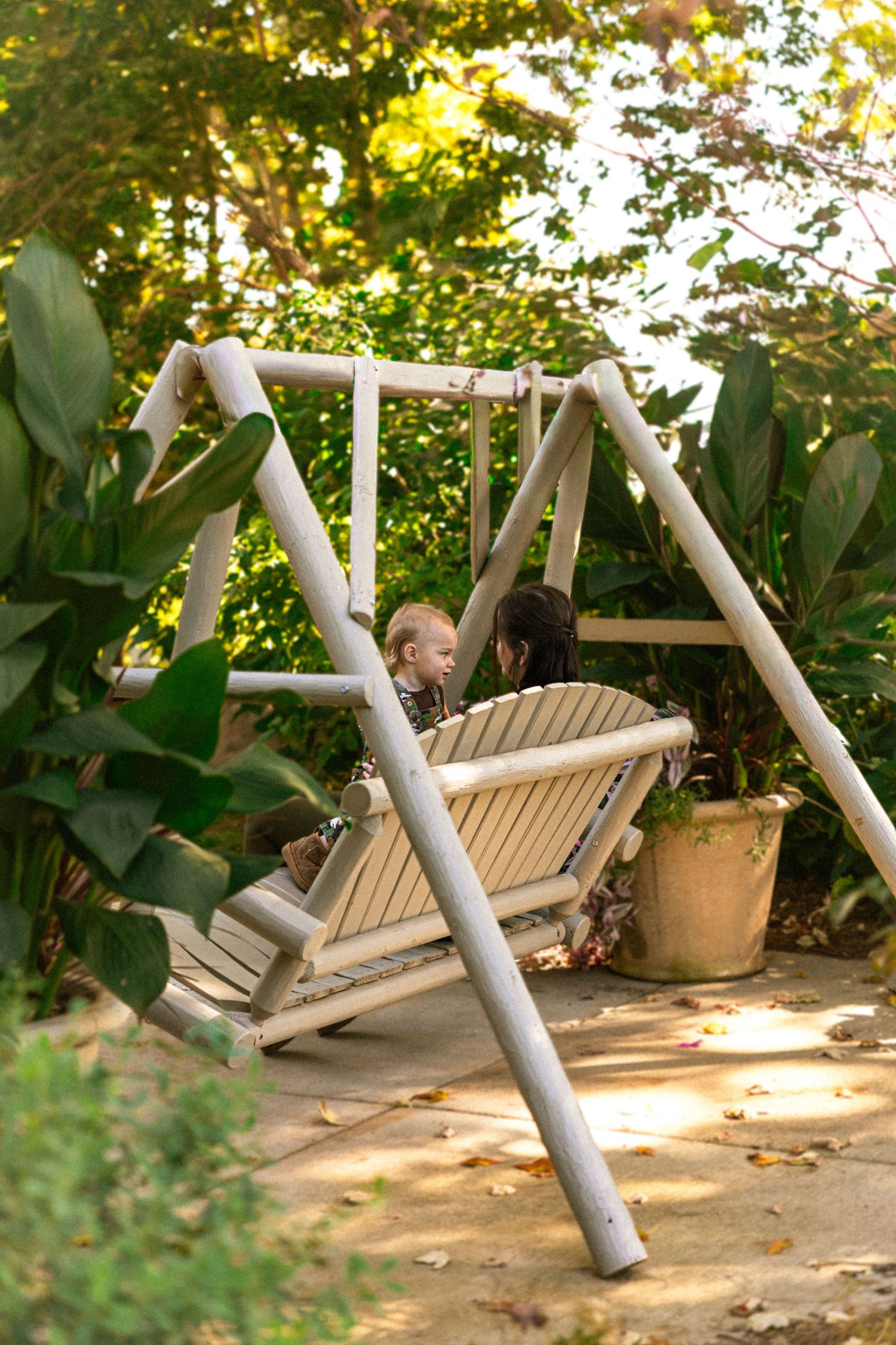 Mother and child sitting together on a wooden porch swing surrounded by lush green garden foliage.