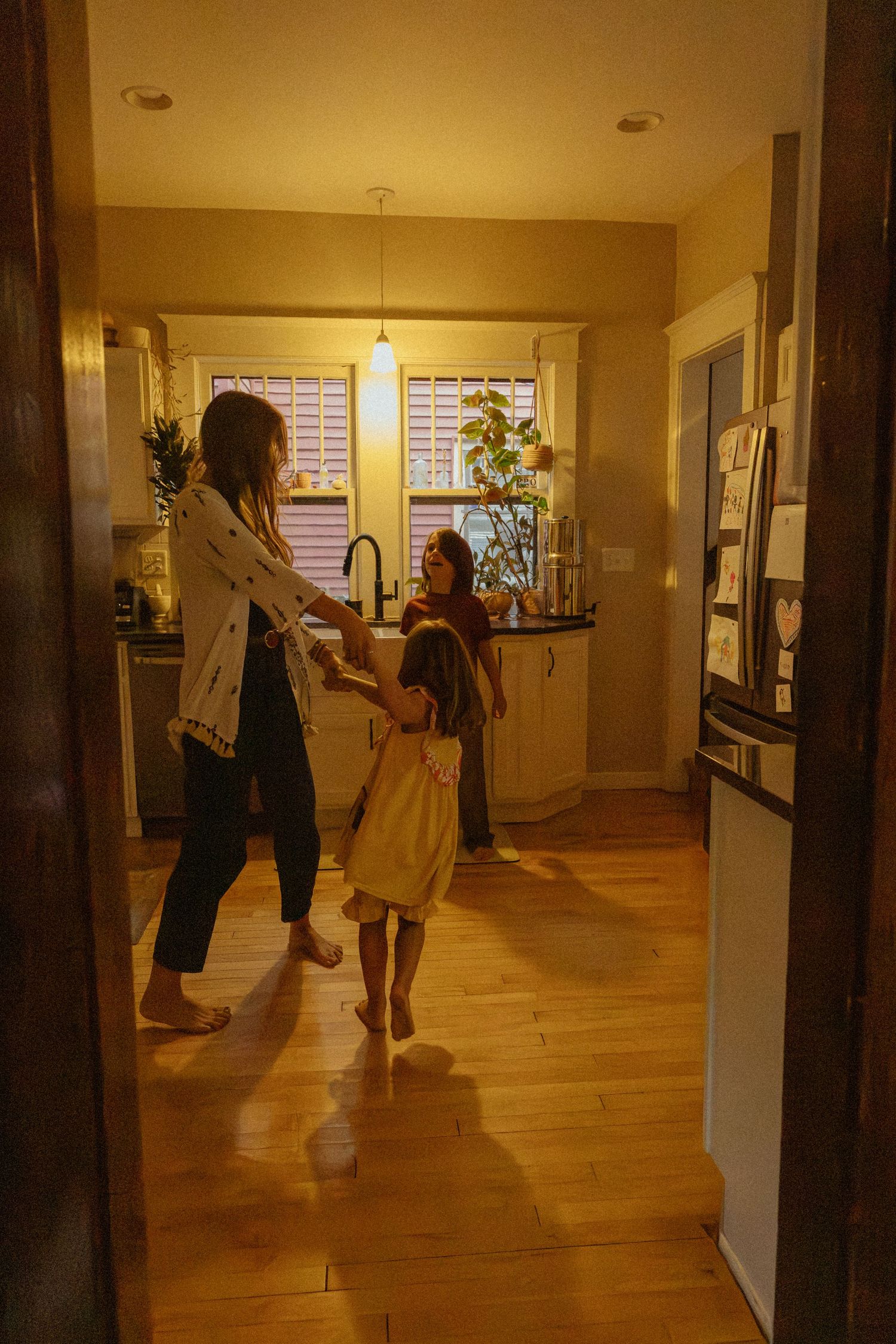 A woman holds hands with a barefoot girl carrying a yellow bag as they dance in a warmly lit kitchen with wood floors.