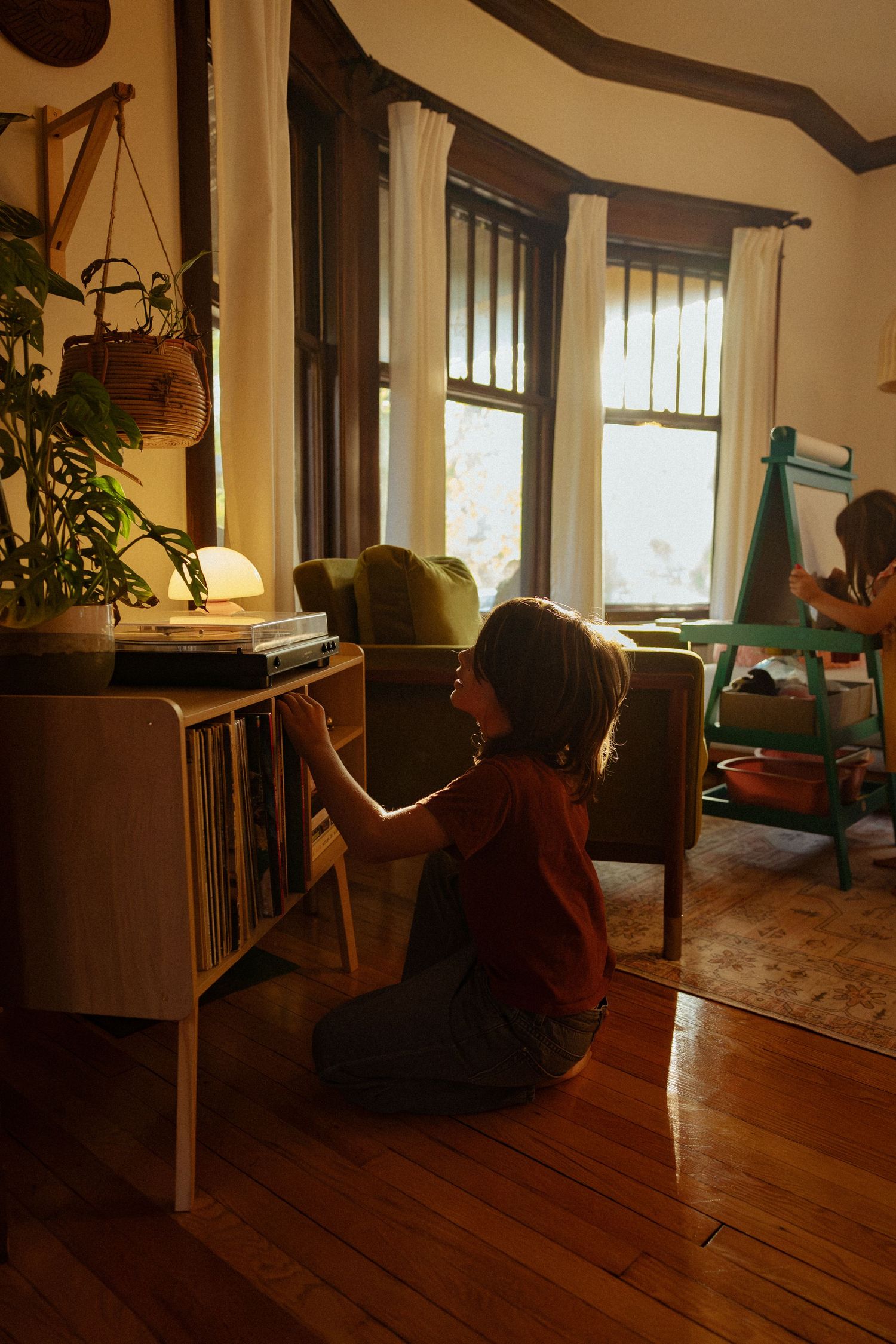 A child kneels on a wood floor flipping through vinyl records on a mid-century shelf in a warm, sunlit living room with houseplants.