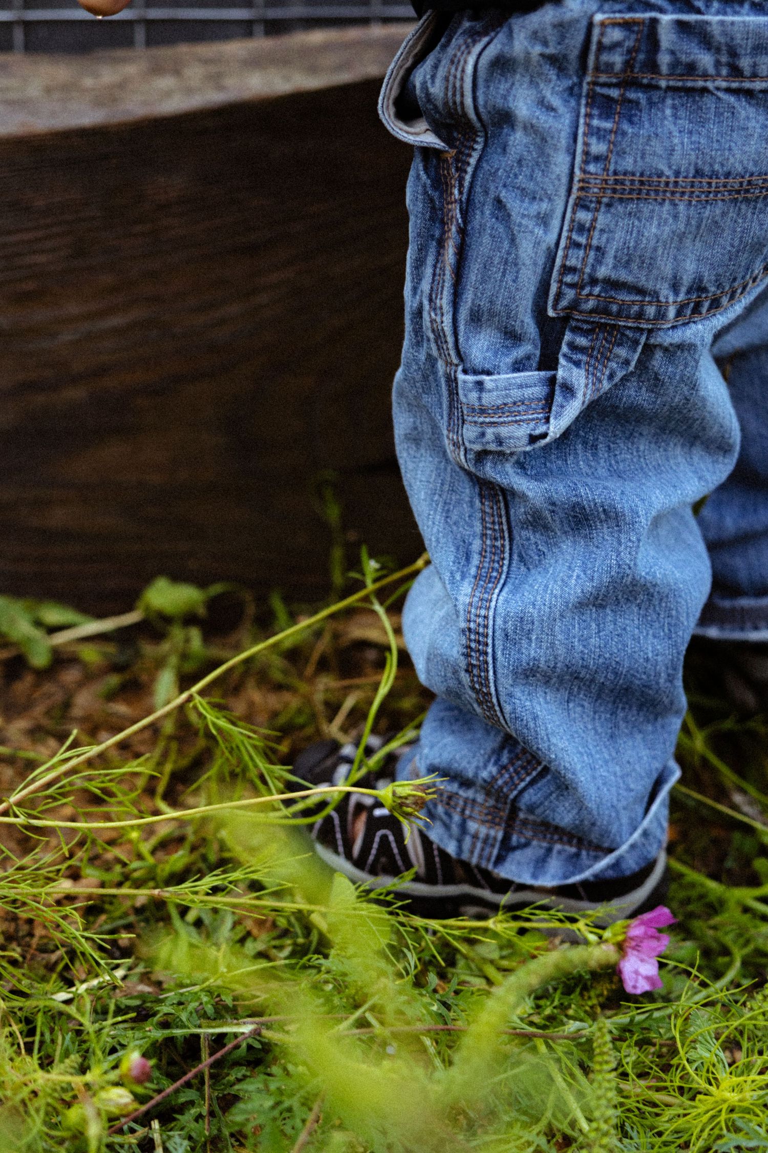 Toddler in blue denim jeans and sneakers standing on green grass near a wooden planter outdoors.