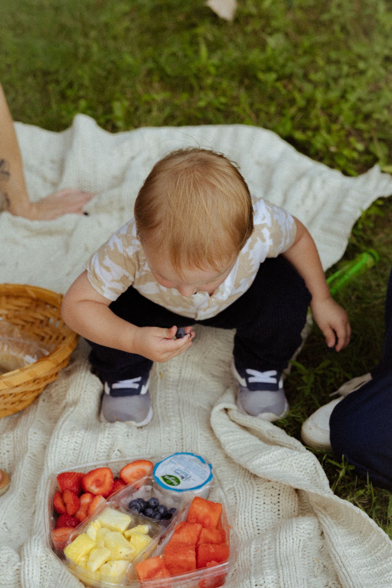 Toddler sitting on a blanket outdoors, exploring a colorful fruit snack container at a picnic.