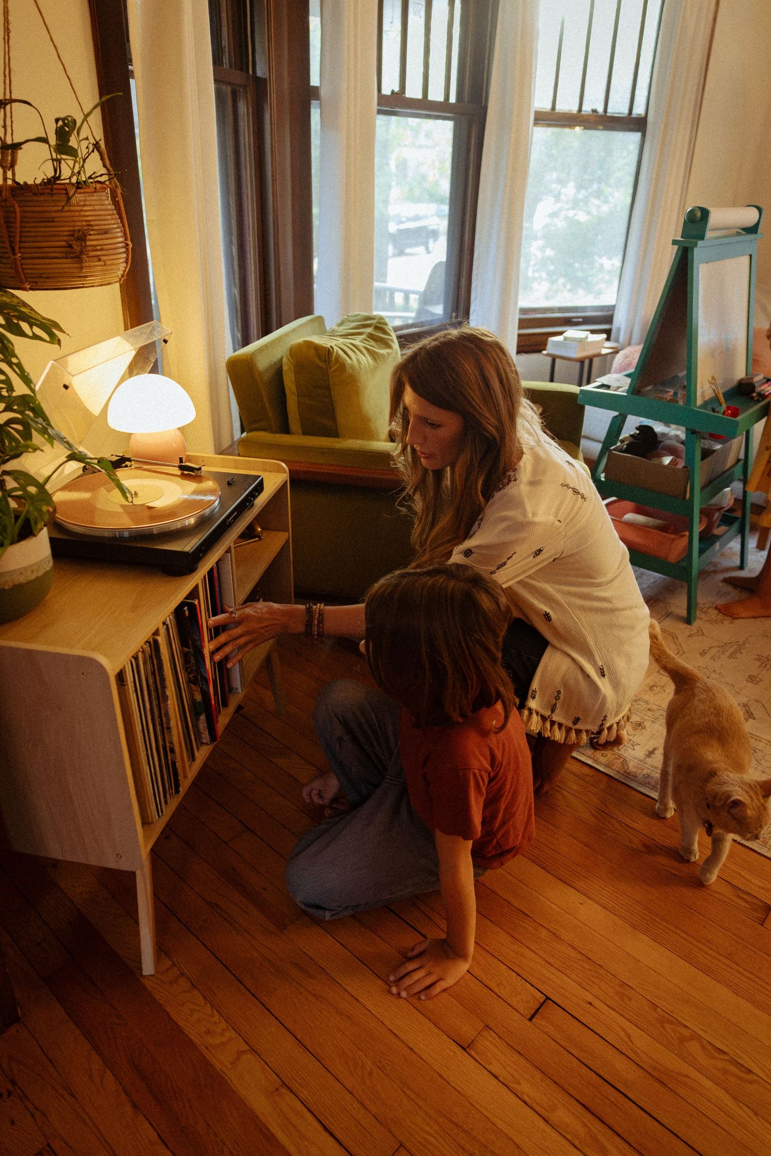 A woman and a child sit on a hardwood floor beside a turntable and record shelf as a cat walks by in a sunlit room.