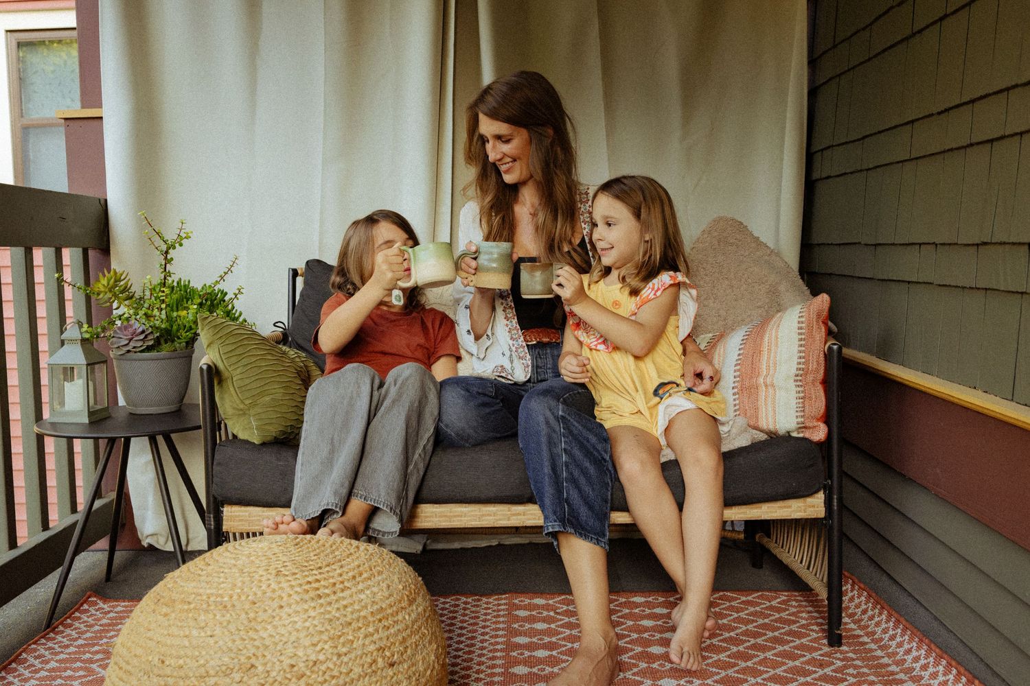 A smiling mother and two young girls sit barefoot on a cushioned porch sofa, sipping from mugs and enjoying a cozy morning together.