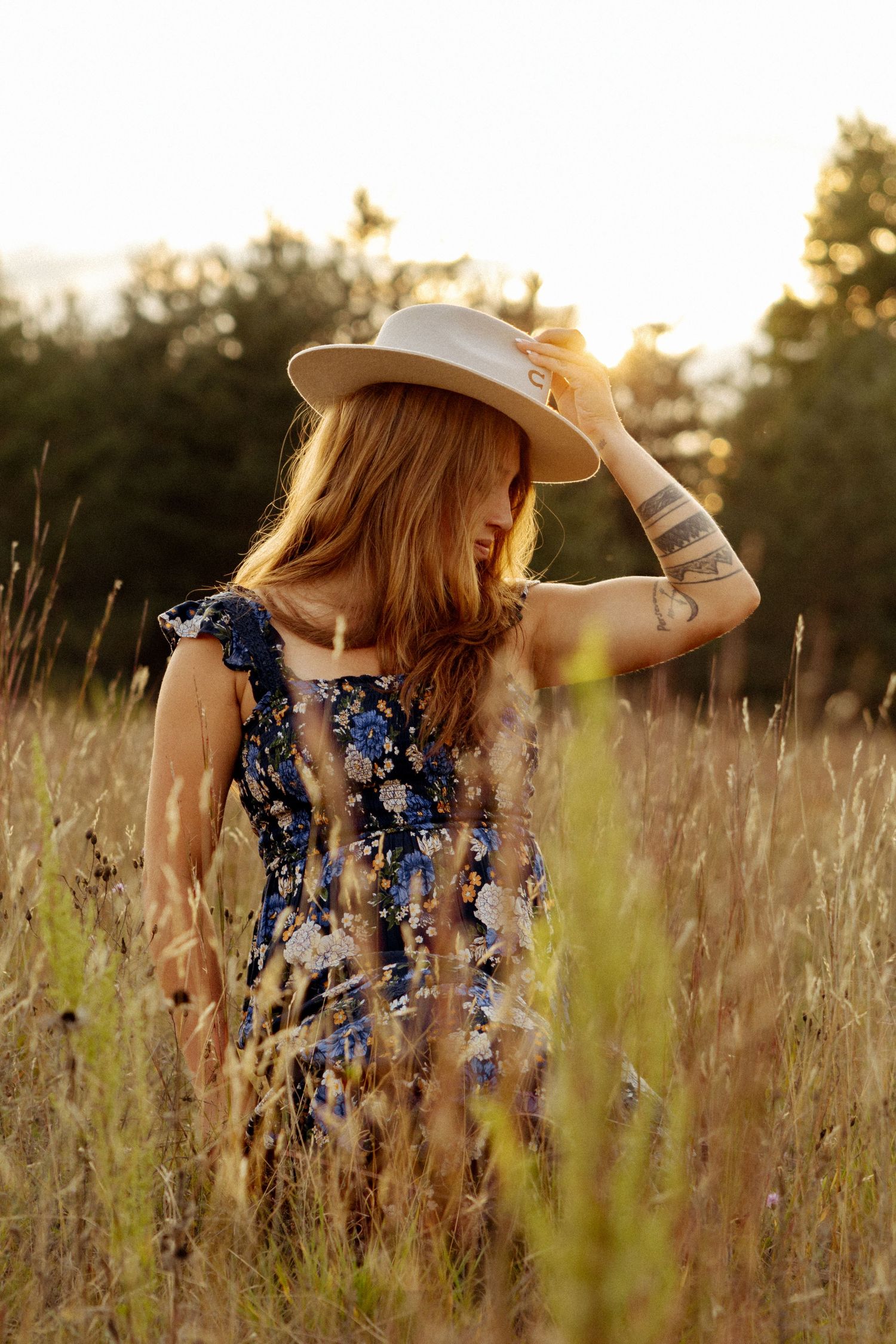 Woman in floral dress and white hat standing in golden field at sunset with tattoos visible.