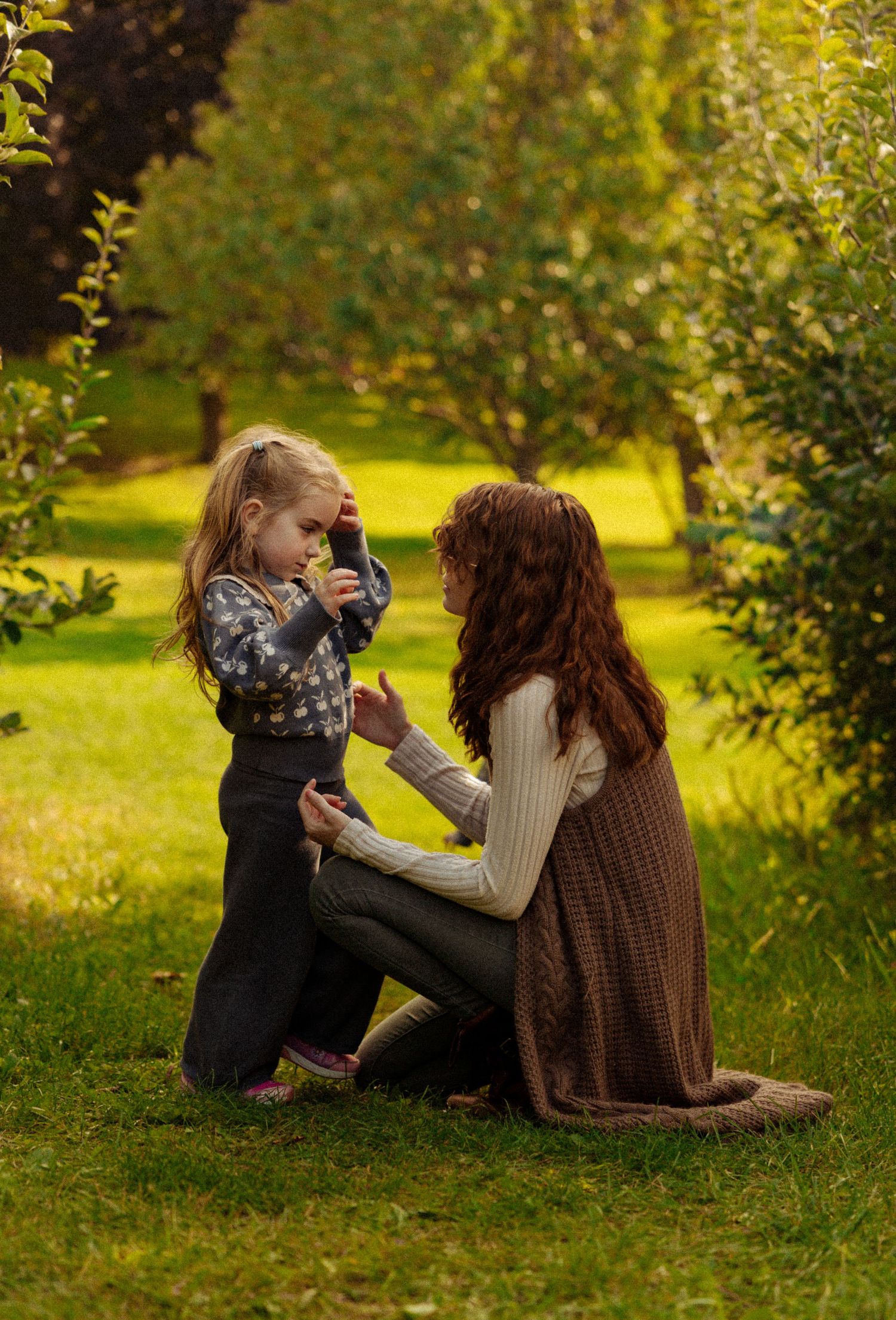Mother kneeling on grass interacting with young daughter in a sunlit green orchard setting.