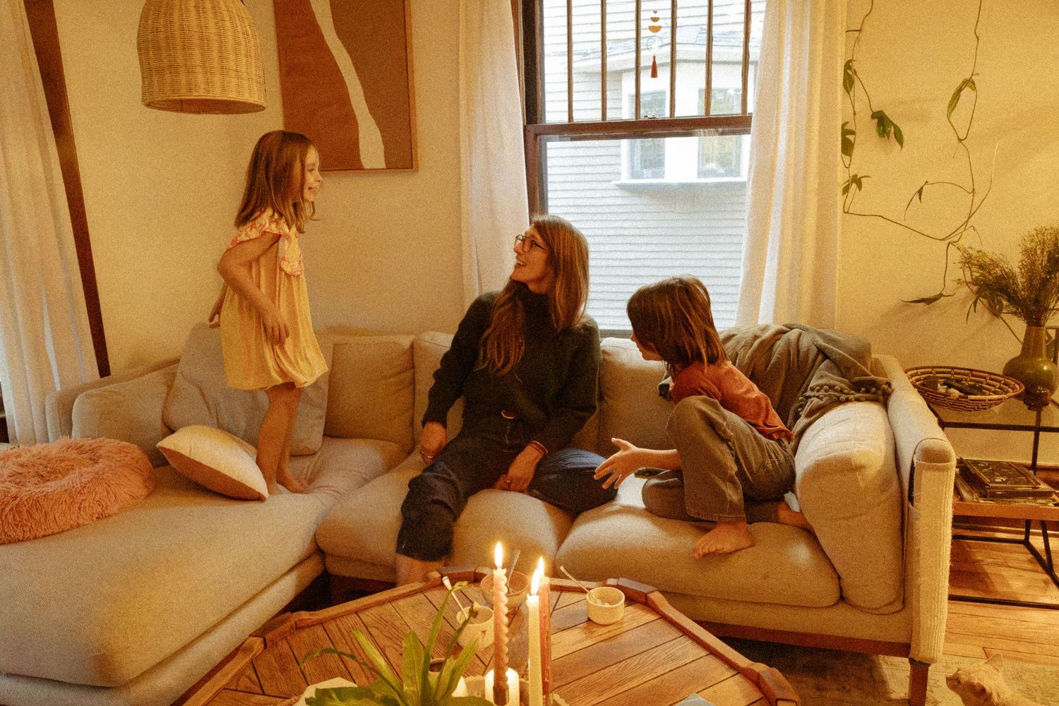 A mother and two young daughters relax together on a cozy cream sofa in a sunlit living room with candles, plants, and boho decor.