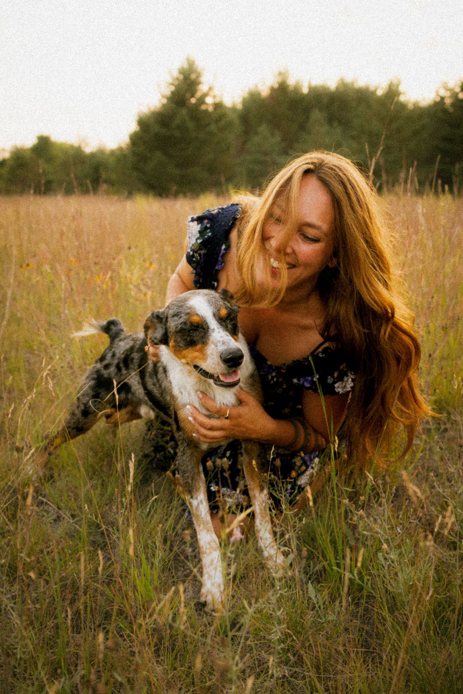 A smiling woman with long red hair hugs a merle dog in a golden field with trees in the background.