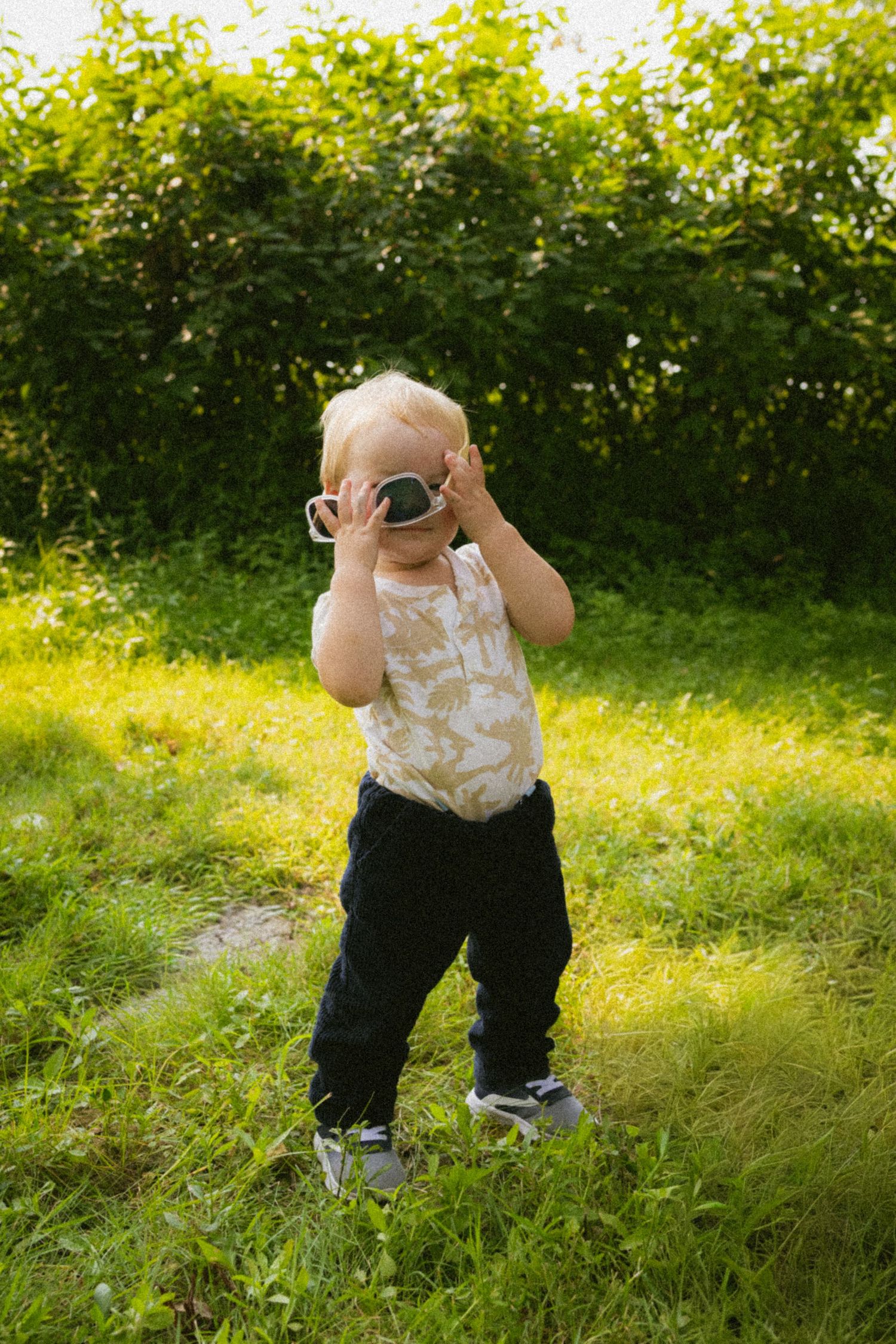 Toddler in floral onesie adjusting oversized sunglasses while standing on grass with green bushes in background.