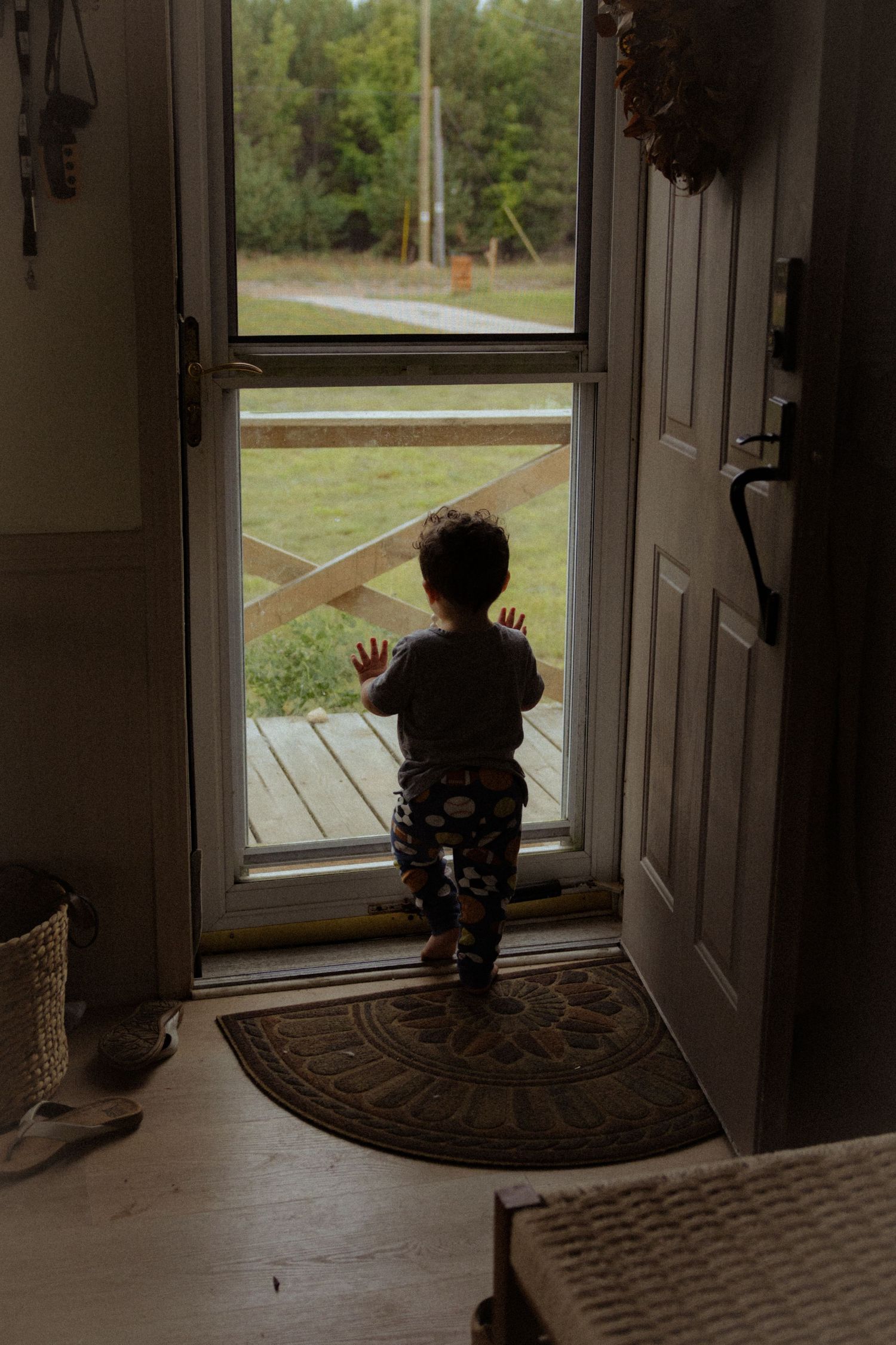 Toddler standing at a glass door looking outside at a green yard and wooden deck.