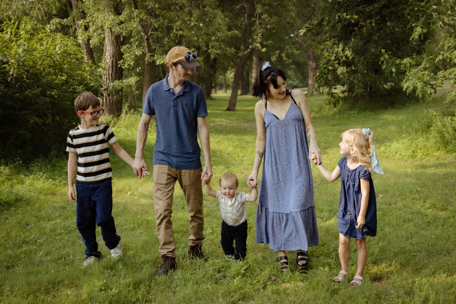 A happy family of five holding hands and walking together on a green grassy lawn in a sunny park.