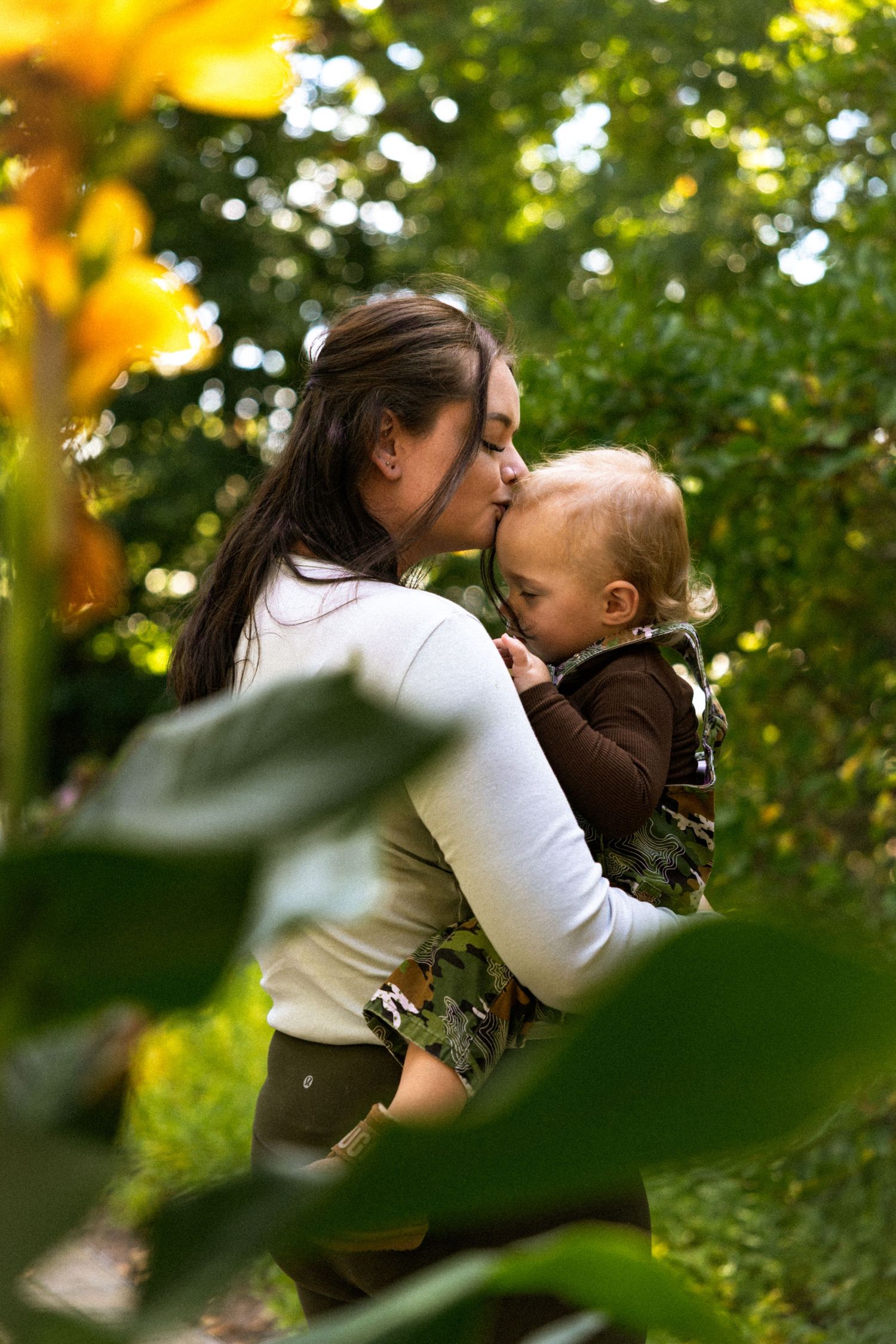Mother kissing her baby outdoors surrounded by lush green trees and yellow sunflowers on a sunny day.