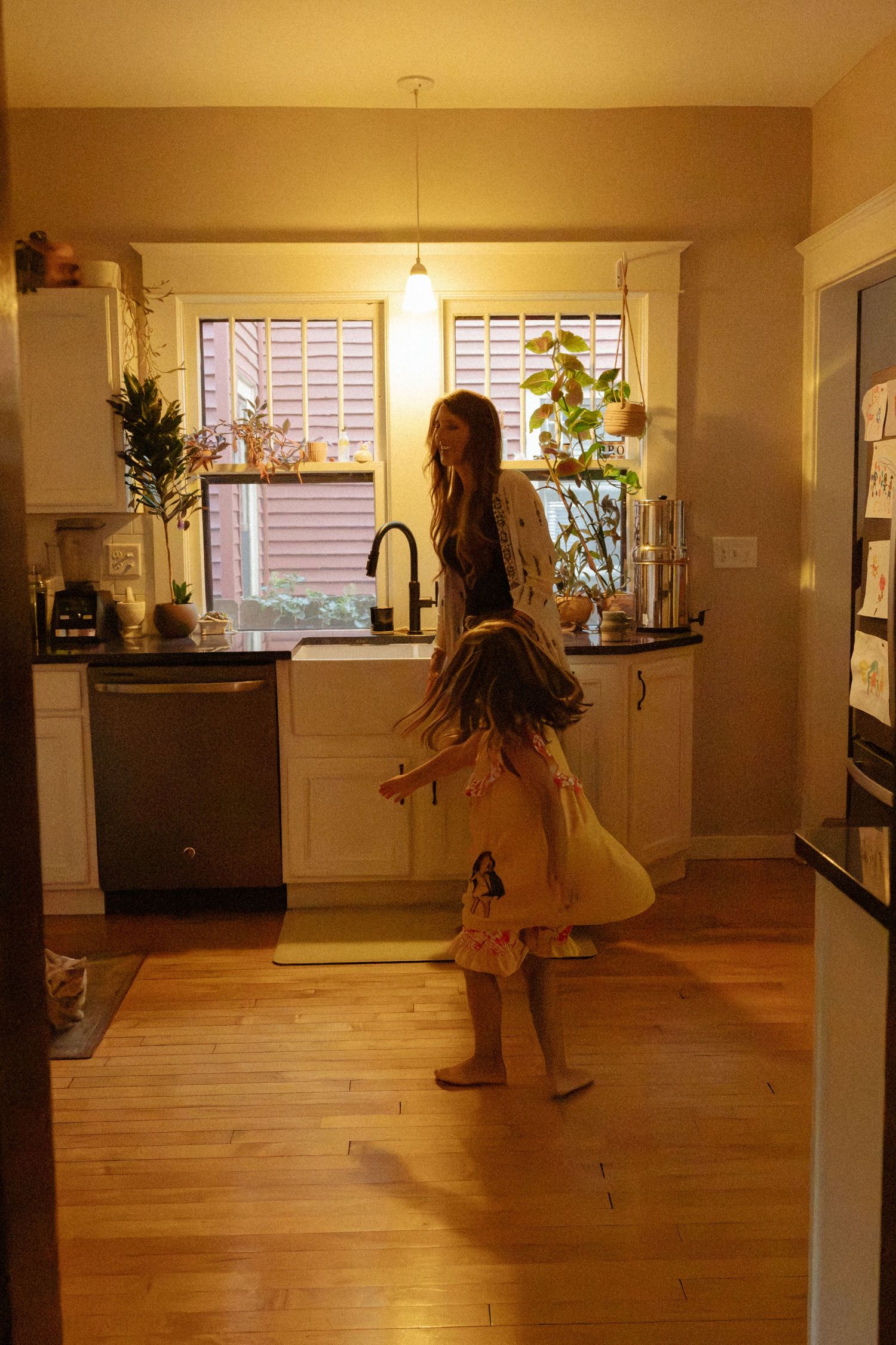 A woman stands at a kitchen sink under a pendant light while a barefoot girl in a pink dress twirls on the wood floor.