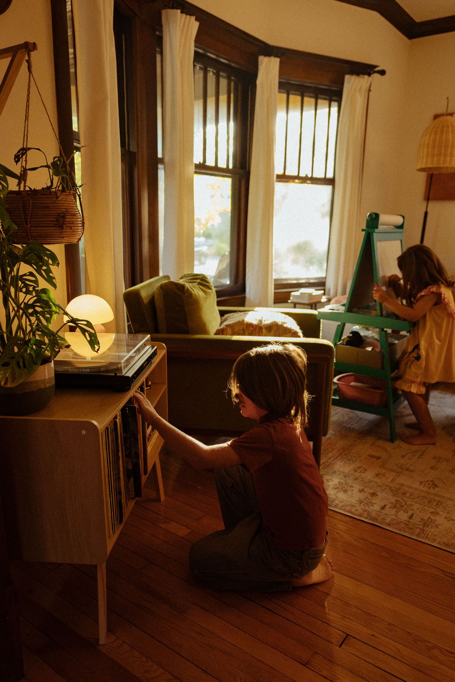Two children play in a sunlit living room with hardwood floors, one browsing a record shelf while the other uses a green easel.