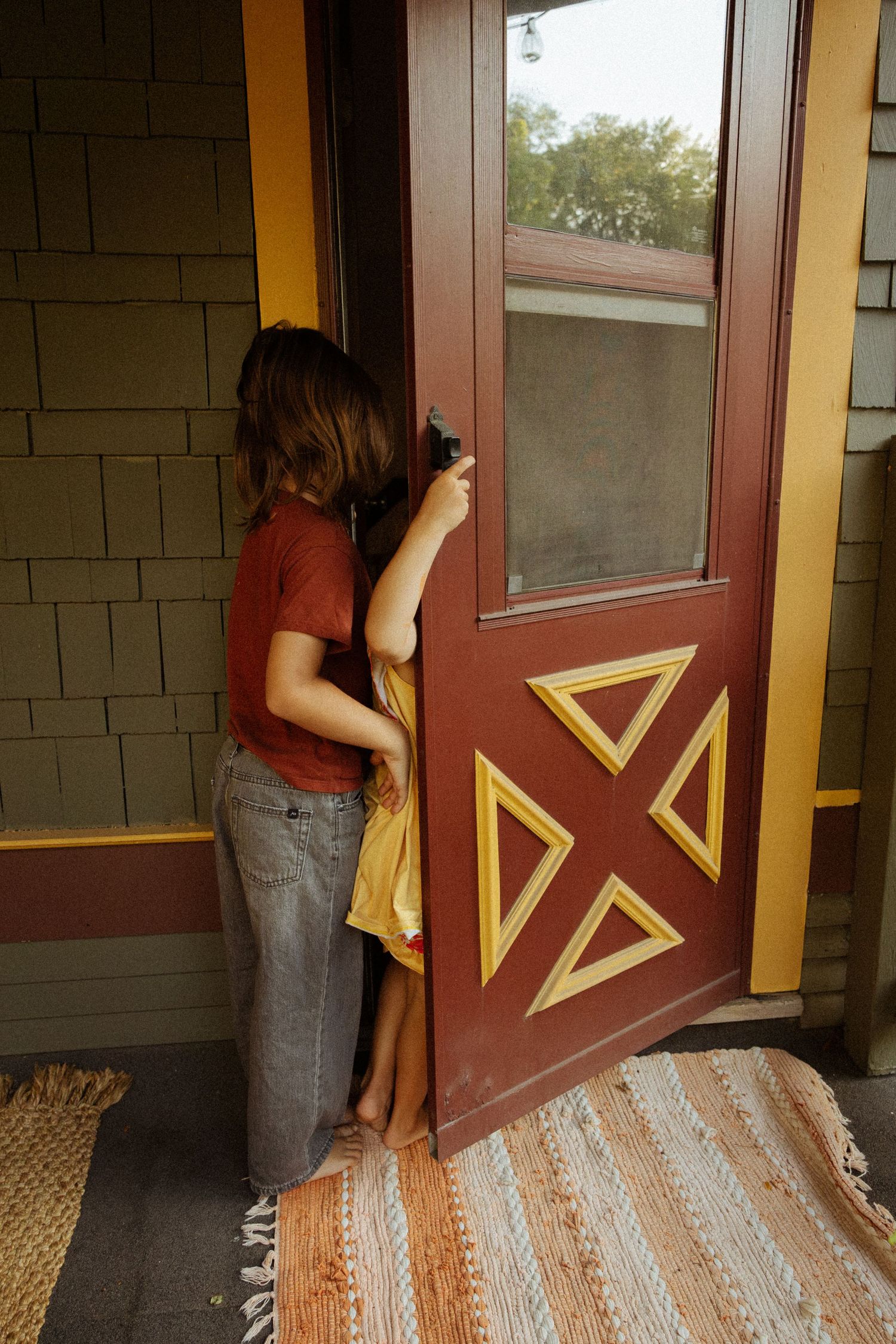 Child in a rust-red shirt and gray pants opens a red wooden door with yellow triangle trim beside a striped woven rug.