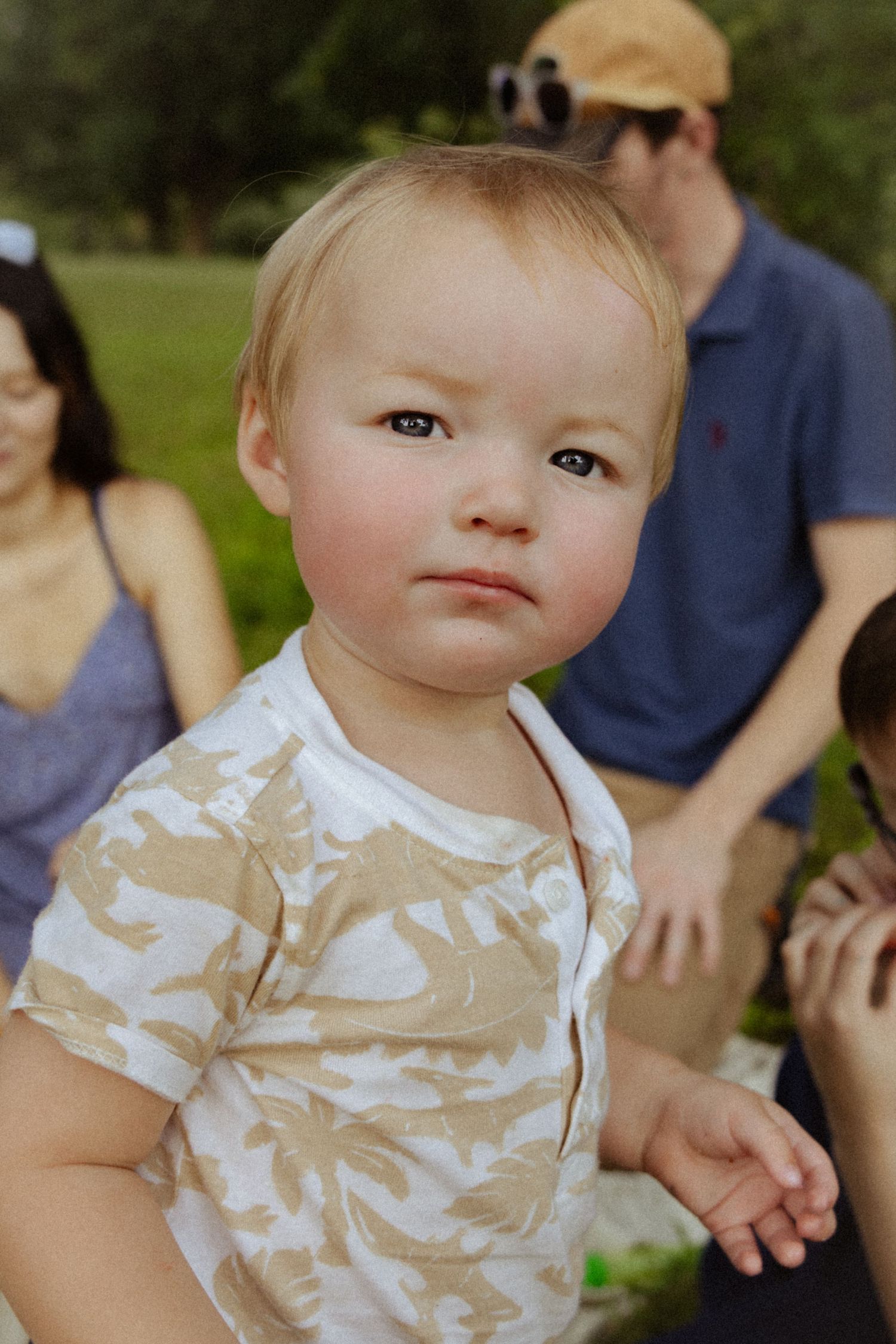 Adorable blonde toddler in camo shirt looks at camera during outdoor family gathering in green park.