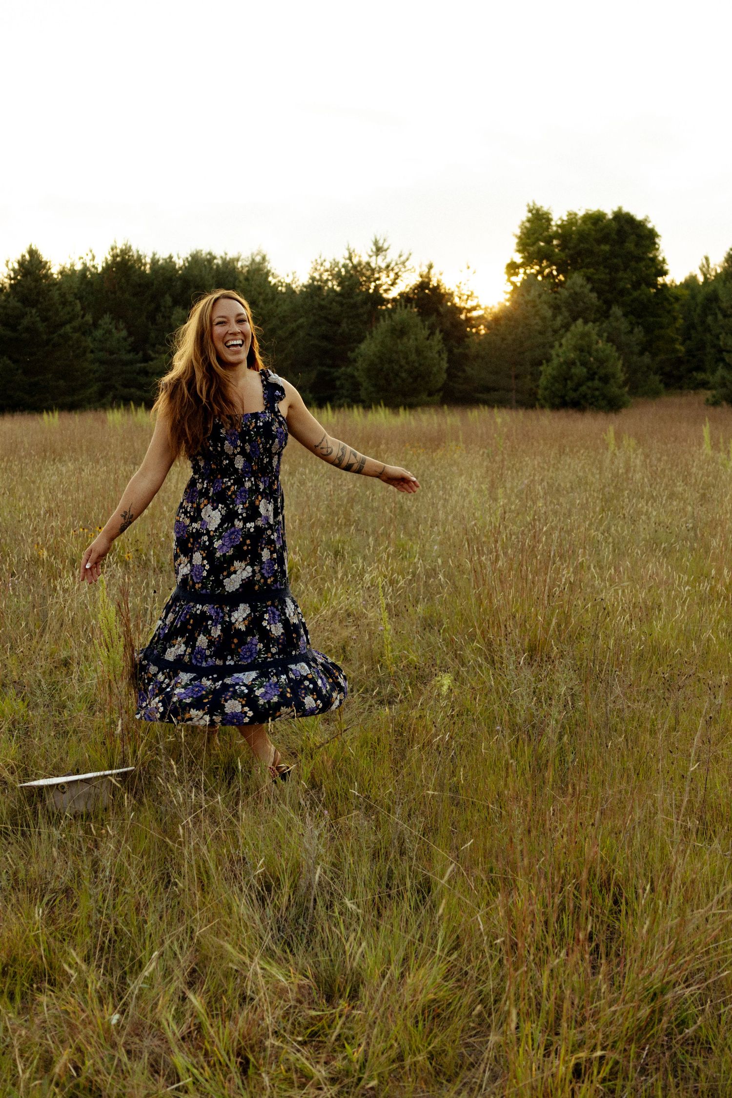 Joyful woman in floral navy dress laughing and dancing in field at sunset.
