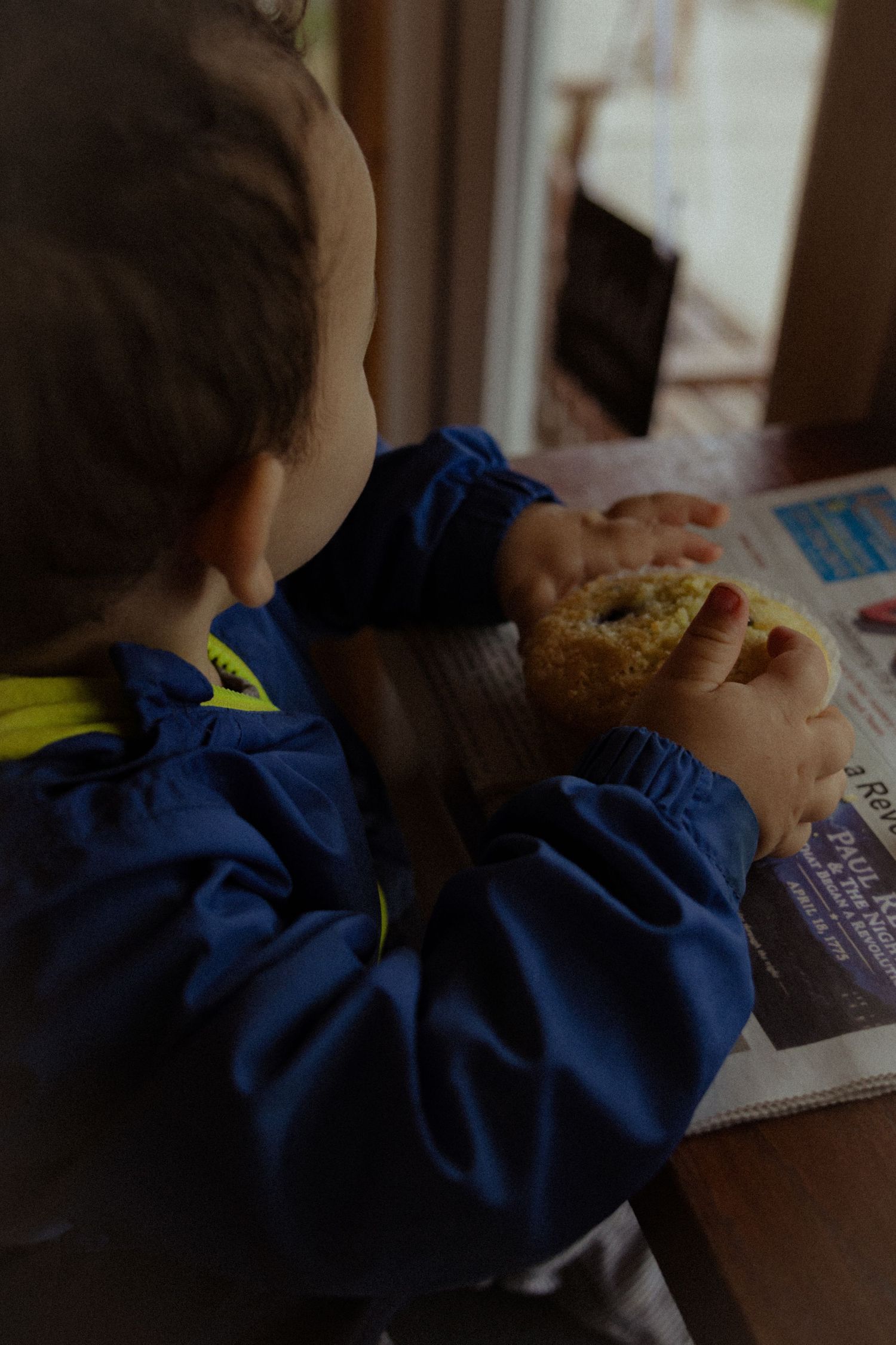Young child in blue jacket eating a snack at a table with newspaper and window in background.