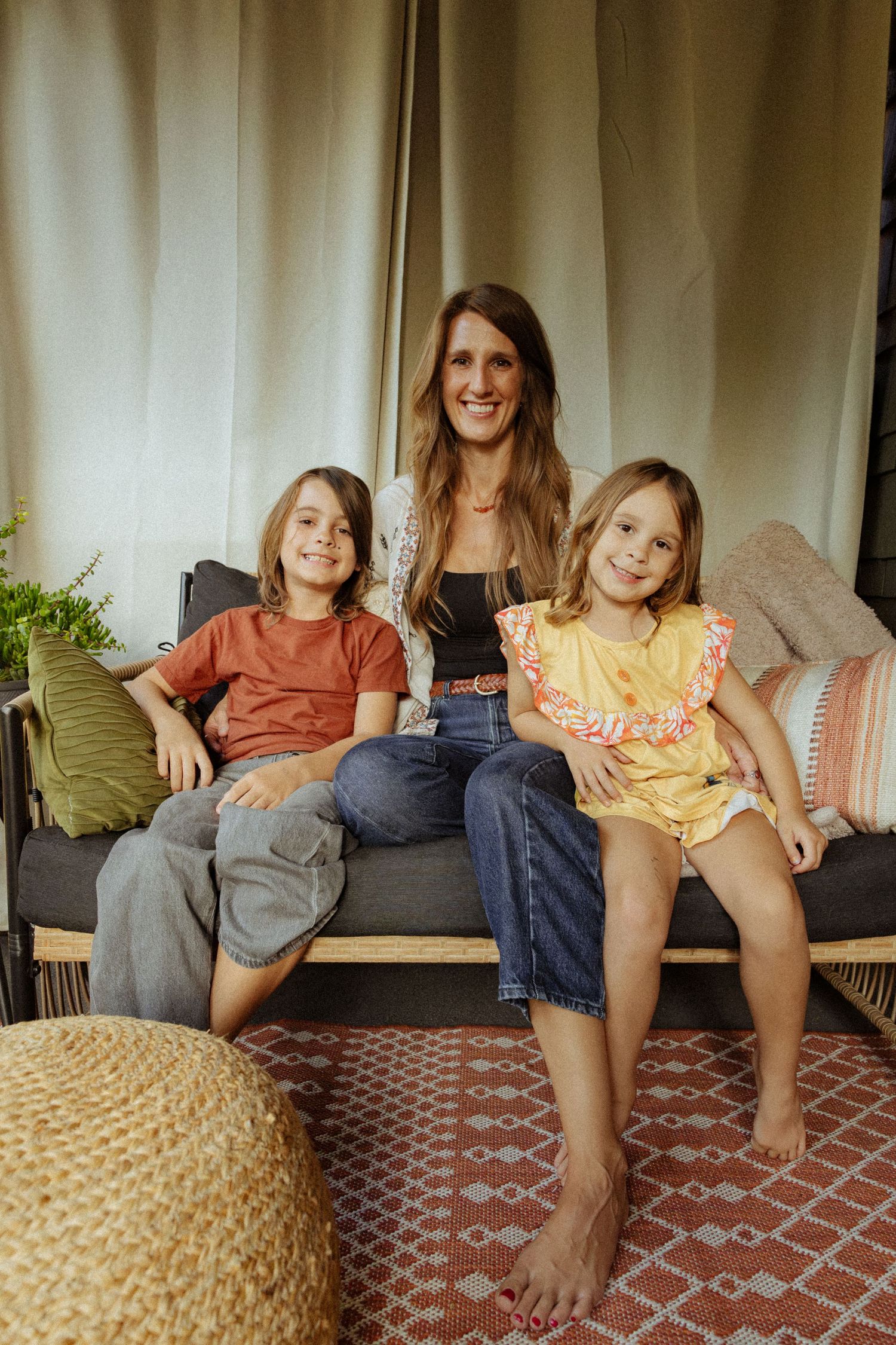 A smiling woman sits on a cushioned outdoor bench between a boy and a girl in a yellow dress, with a red patterned rug below.