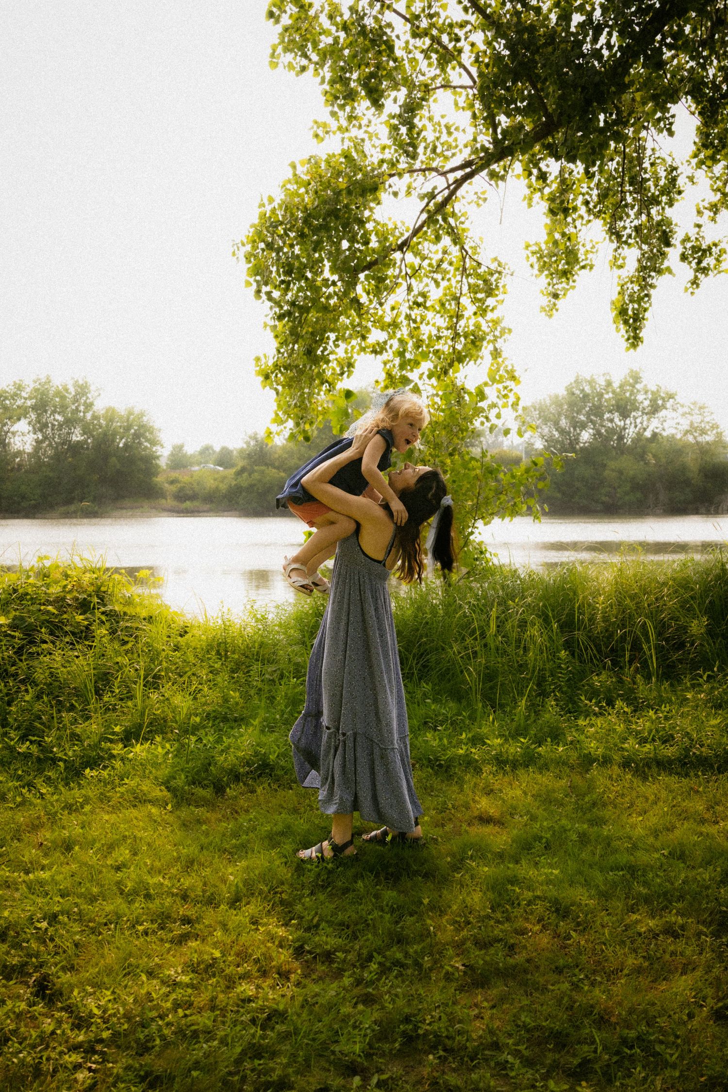 A mother lifts her young child joyfully outdoors near a serene lake surrounded by lush green trees and grass.