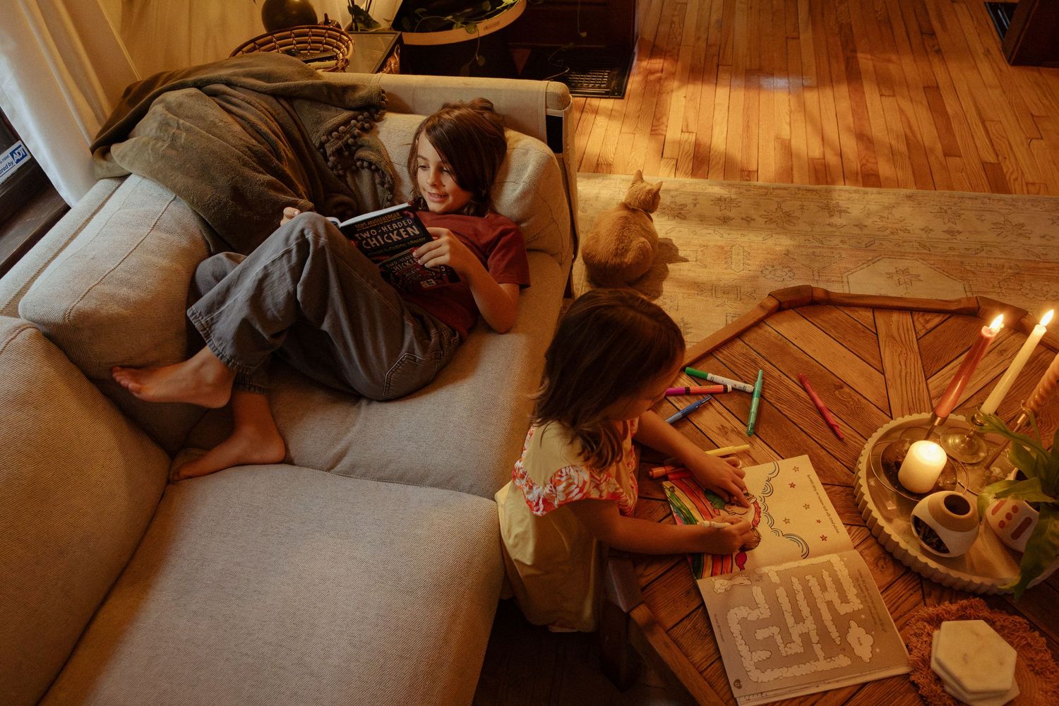 Two children relax in a cozy living room, one reading a book on a gray sofa while the other colors at a wooden coffee table with candles.