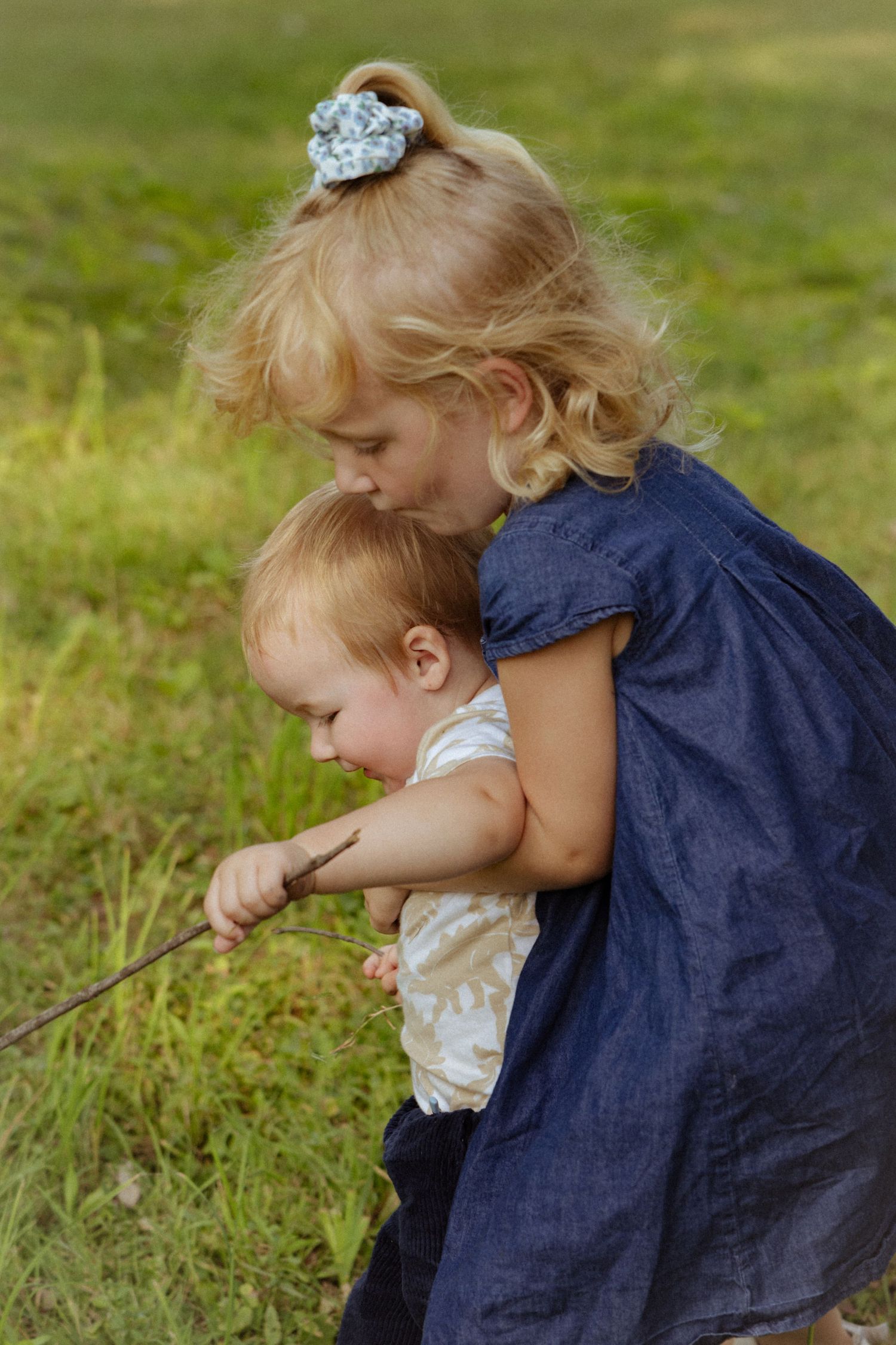 Older blonde girl in denim dress hugging toddler sibling while playing with a stick outdoors in green grass.