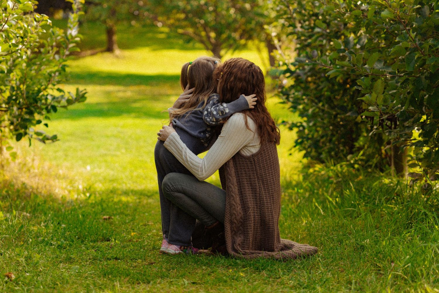 Two people sharing a warm embrace in a lush green garden surrounded by trees on a sunny day.