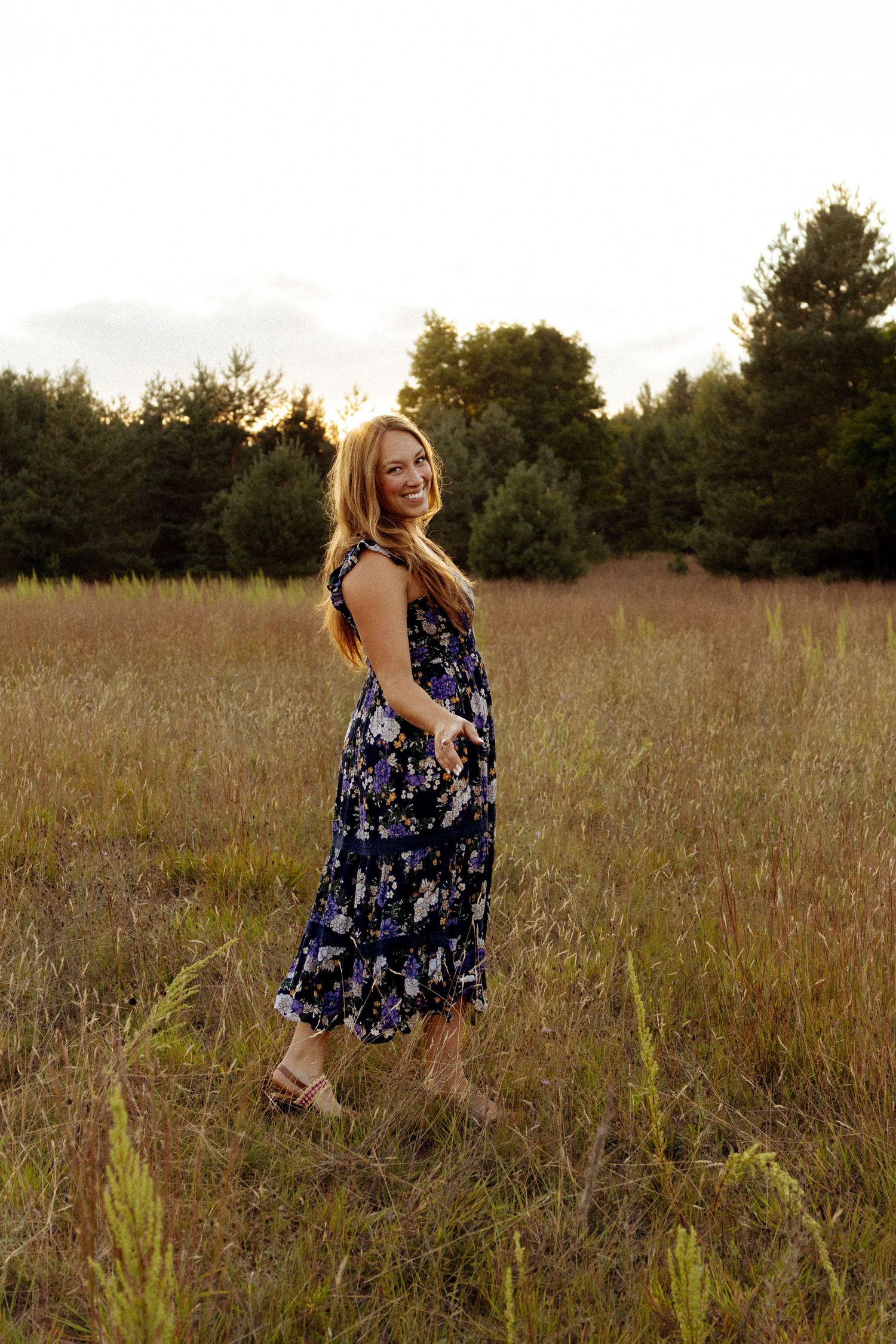 Woman in floral navy dress smiling in golden field at sunset.