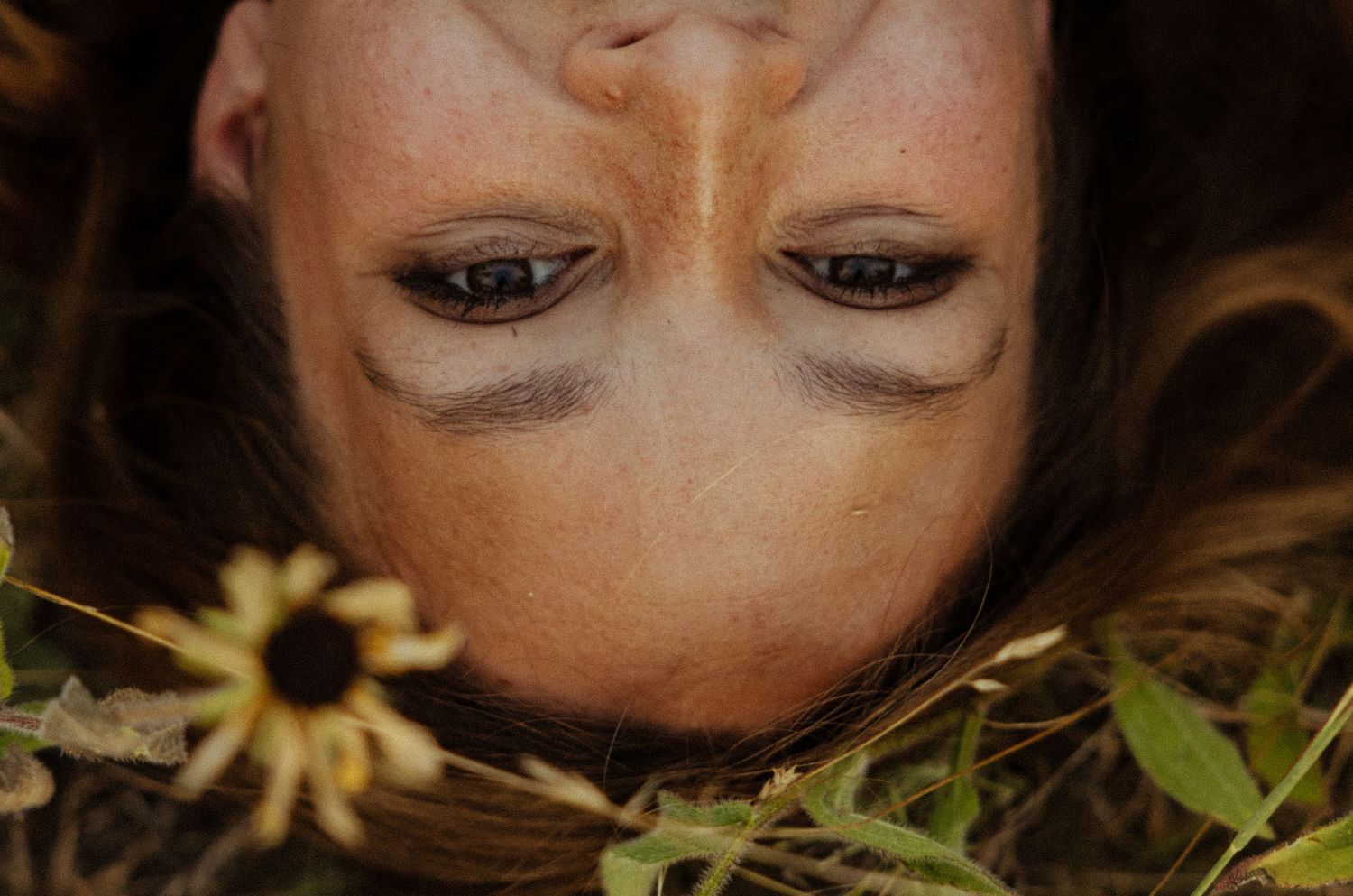 Close-up of a woman lying upside down among wildflowers and green foliage, with freckles and dark eye makeup.